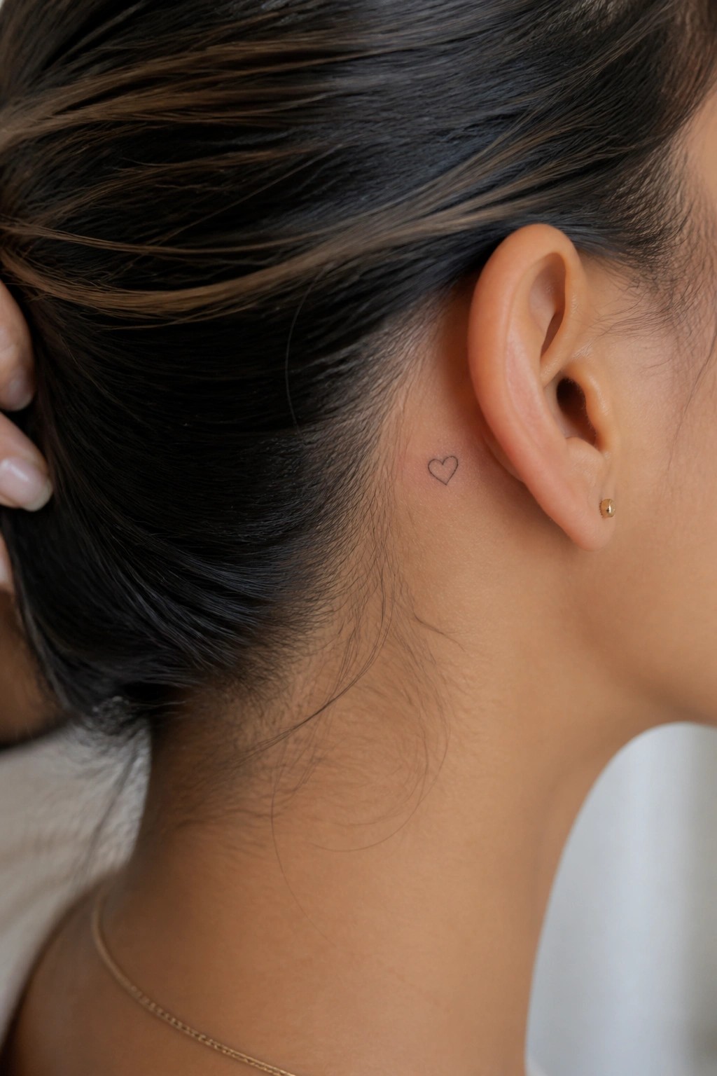 Woman's neck with small heart tattoo behind ear, gold stud earring, and thin necklace.