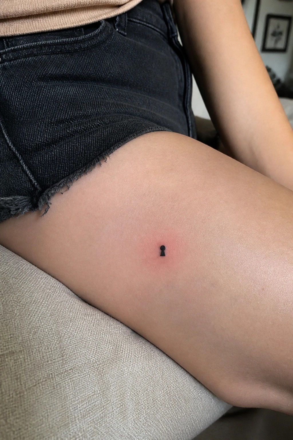 Small black keyhole tattoo on the upper thigh near the hem of black denim shorts