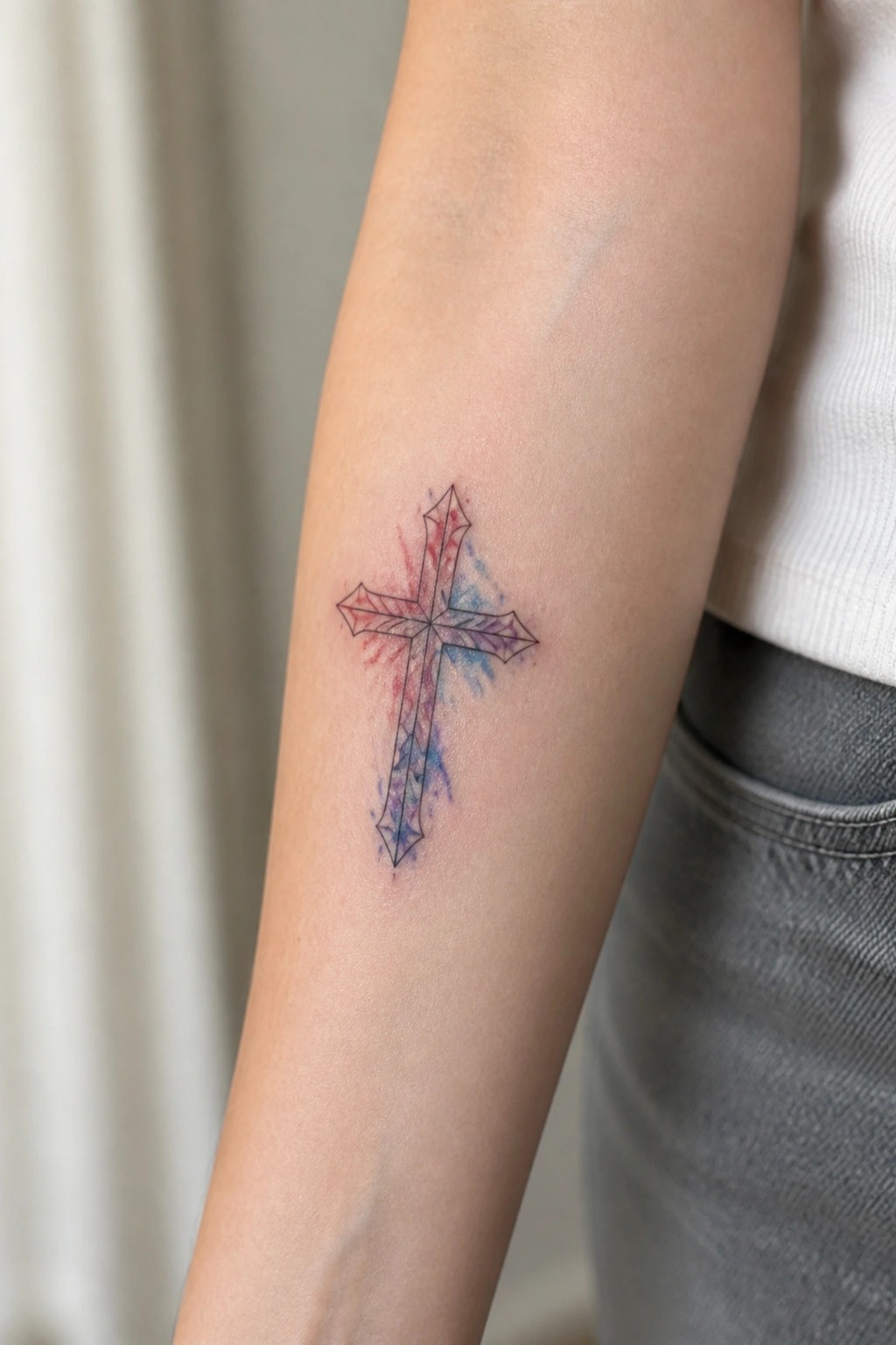 Ornate cross tattoo with red, purple, and blue watercolor splashes on forearm.