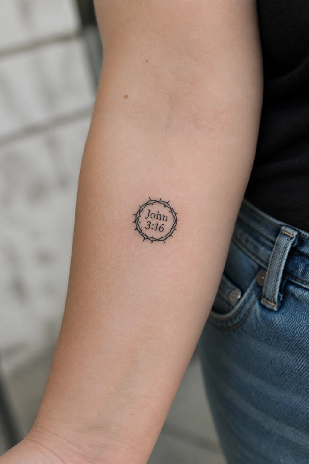 Black ink thorny crown tattoo with John 3:16 on upper arm near blue jeans