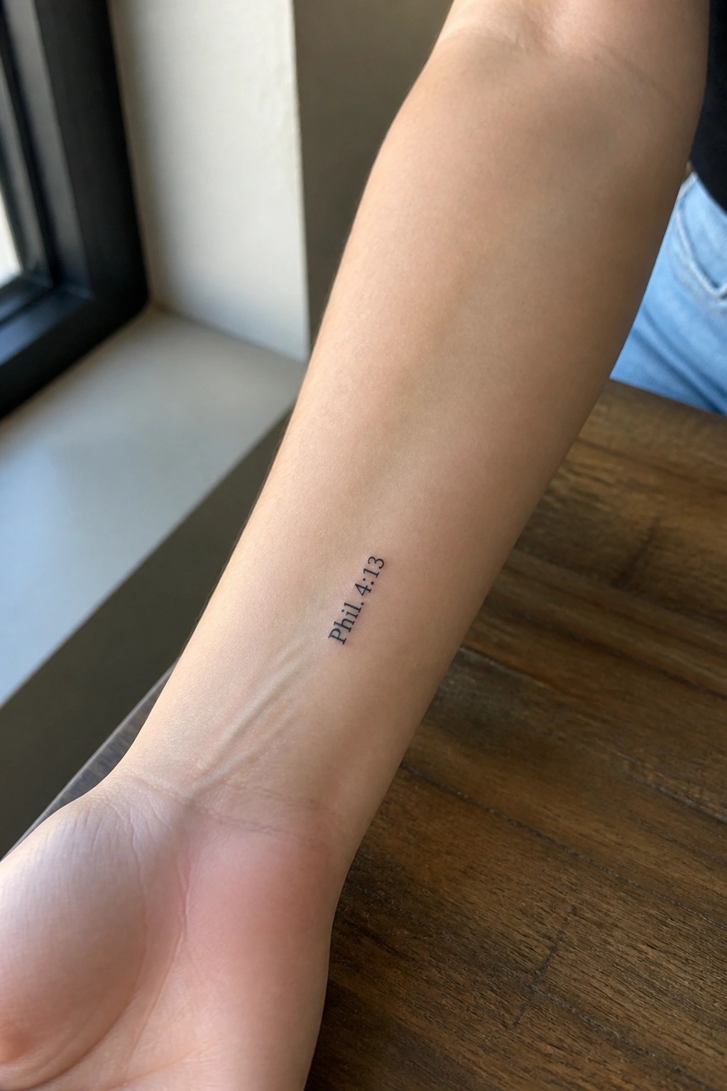 Small black text tattoo of Phil. 4:13 on the inner forearm in a minimalist font.