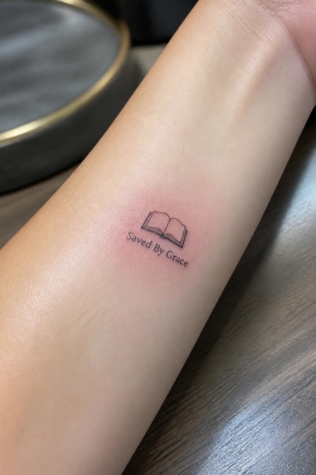 Small open book tattoo with saved by grace text on inner forearm in minimalist line style