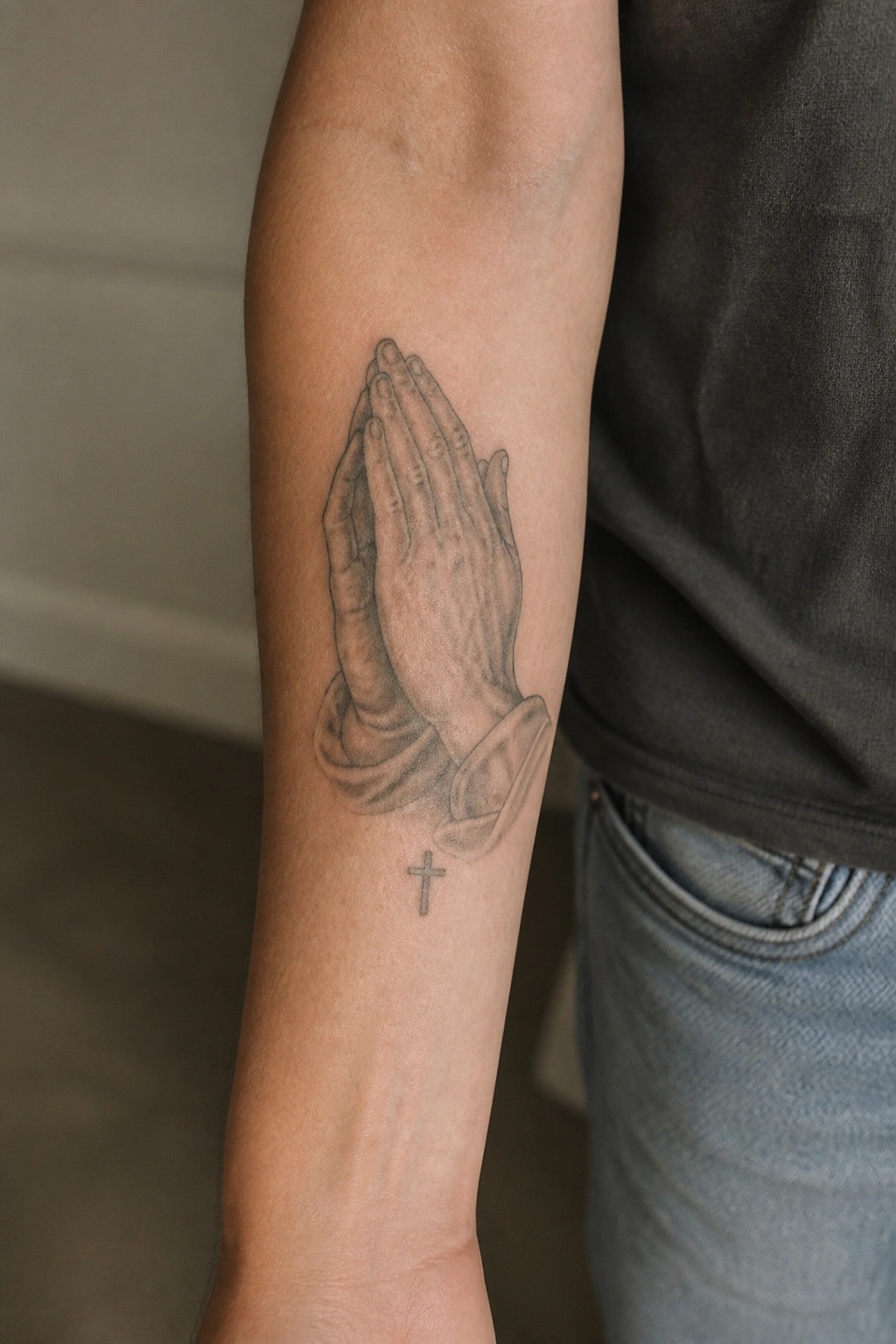 Praying hands tattoo with small cross on forearm, dark shirt and blue jeans visible.