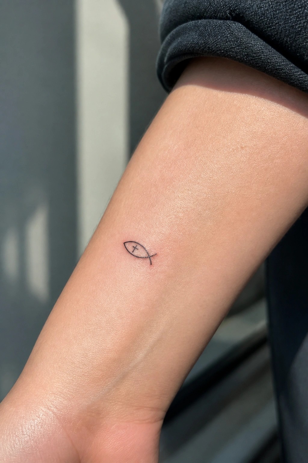 Small black line tattoo of a fish symbol with cross on inner forearm skin