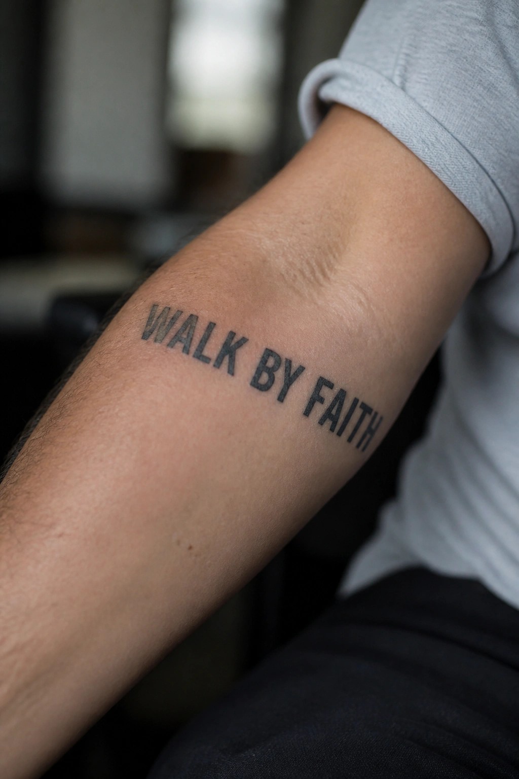 Forearm tattoo spelling 'WALK BY FAITH' beside rolled gray t-shirt sleeve