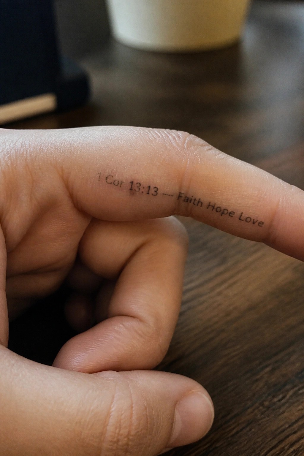 Close up of a small text tattoo on the side of a finger that reads 1 Cor 13:13 followed by the words faith hope love.