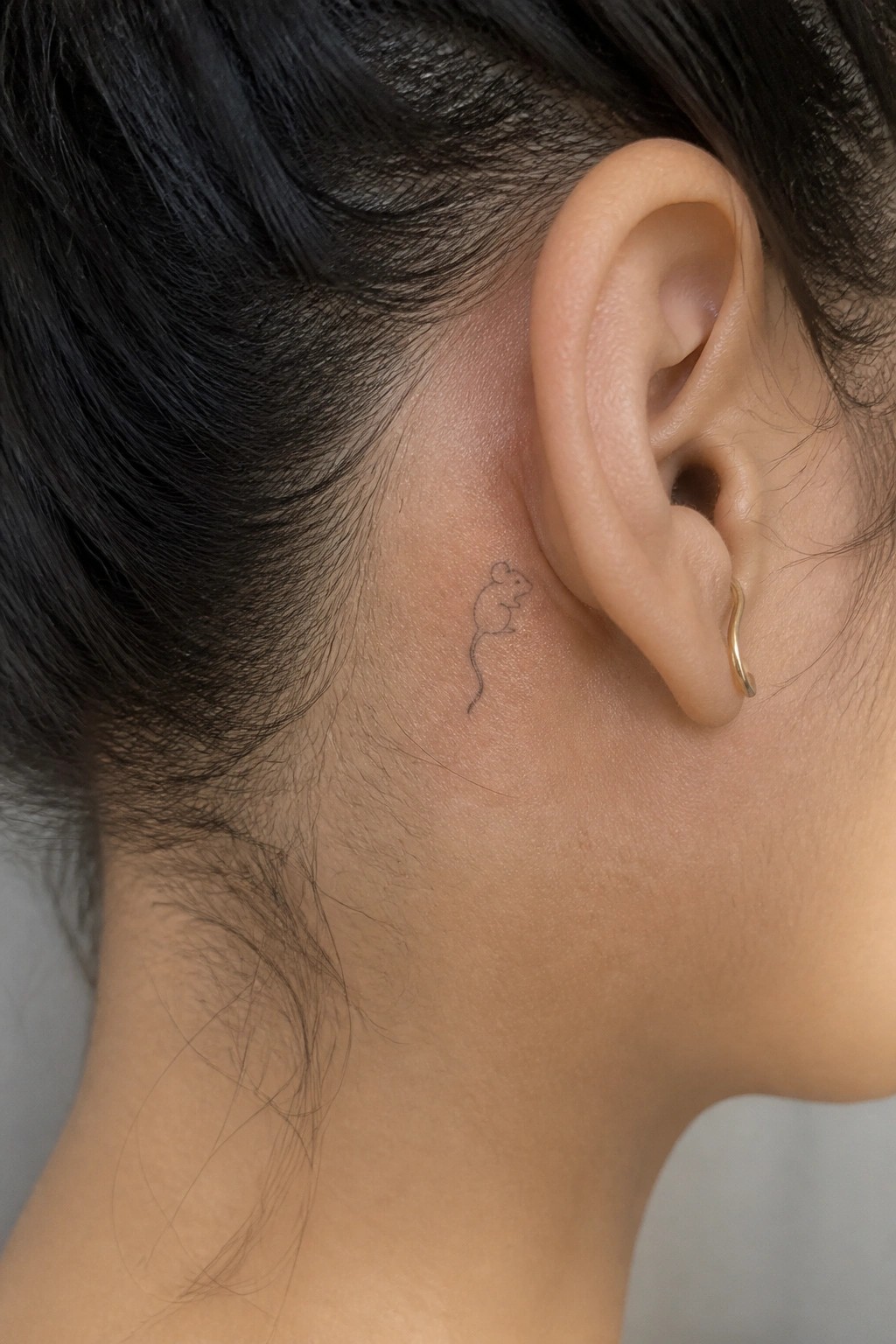 Small mouse tattoo behind ear on dark hair with gold earring