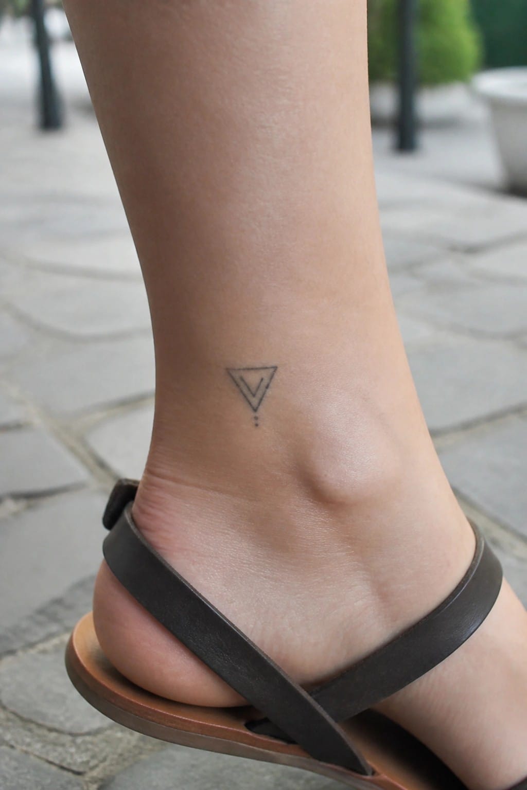 Ankle with small geometric triangle tattoo above brown leather sandal on pavement