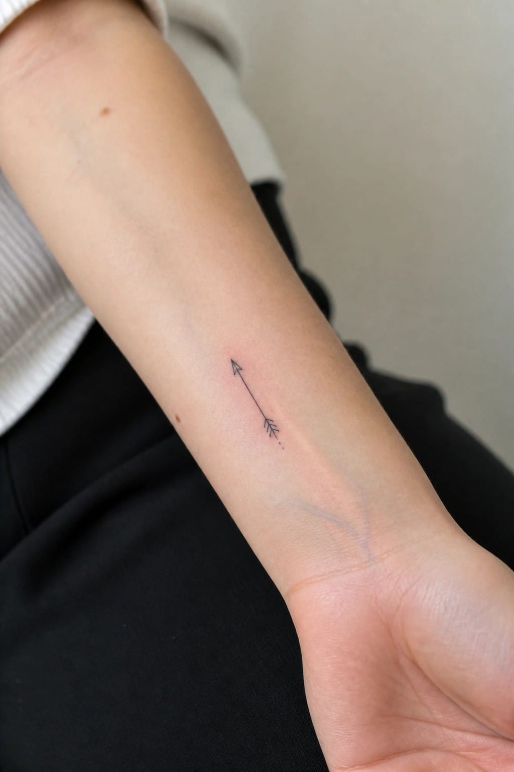 Small black arrow tattoo pointing upward on light-skinned forearm in black clothing