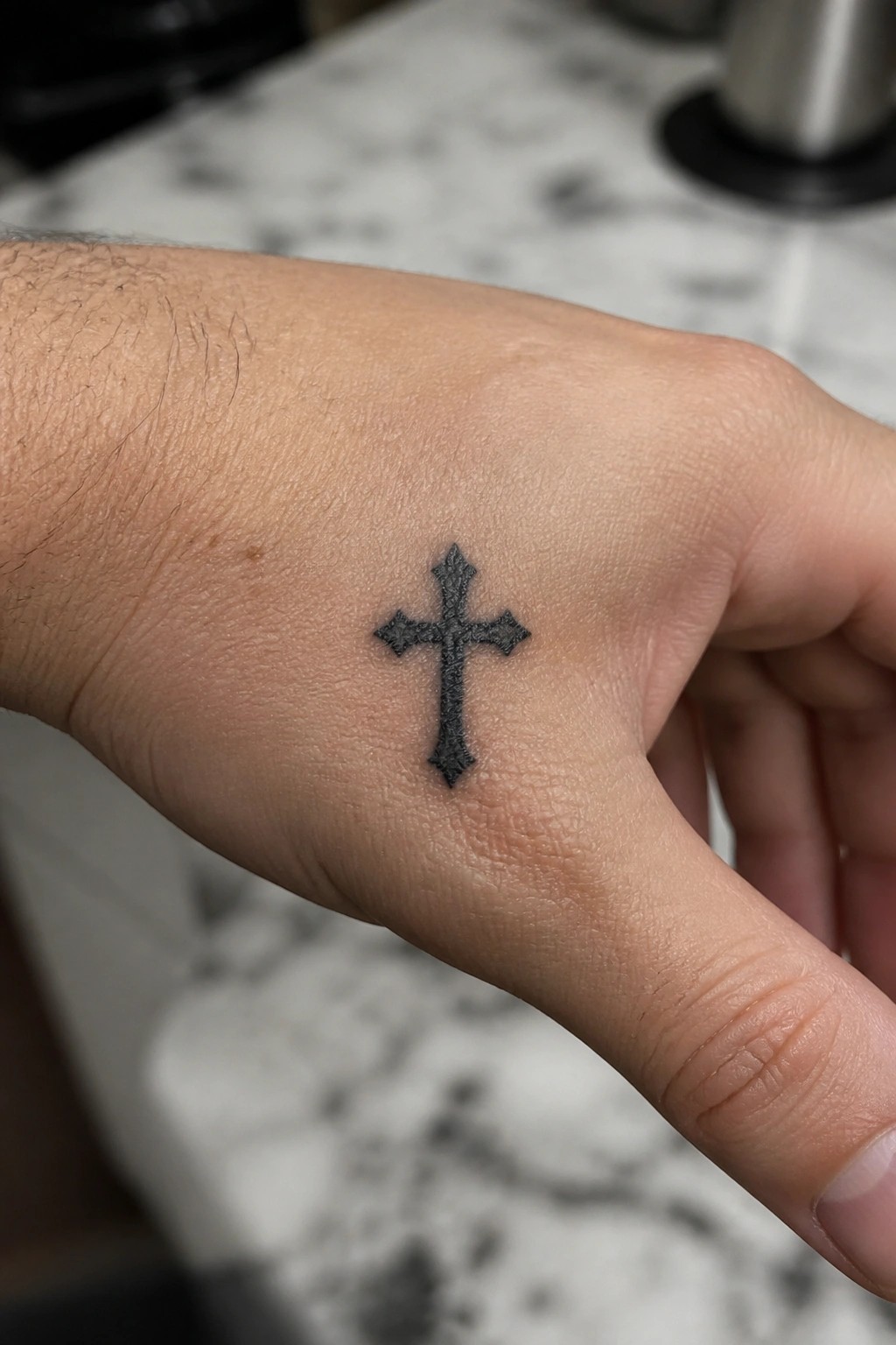 Ornate black cross tattoo on back of light-skinned hand over marble surface