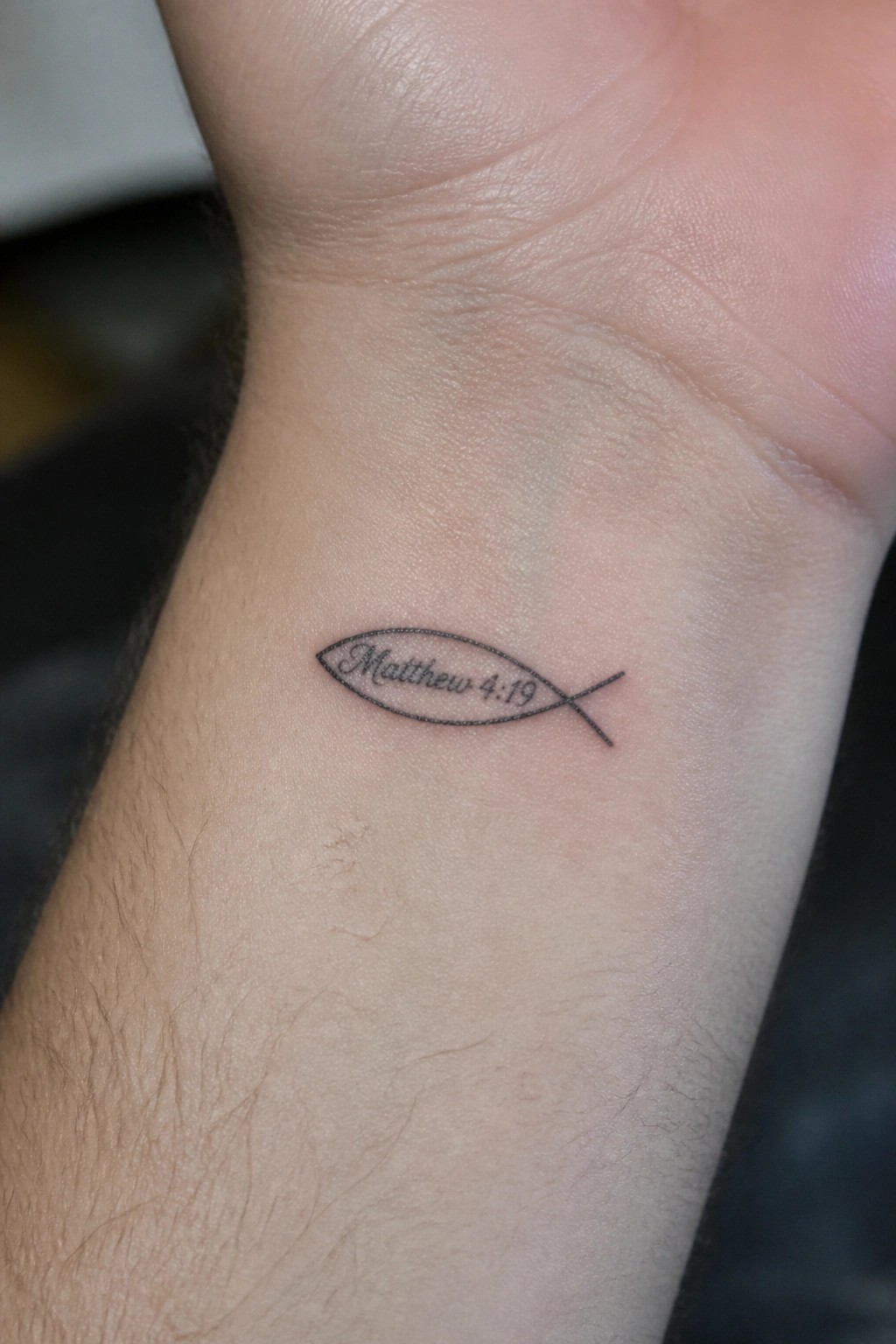 Small fish tattoo on the inner wrist with Matthew 4:19 written inside the outline