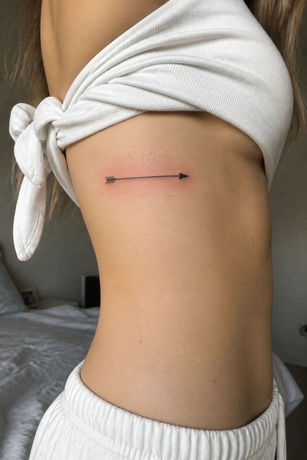 Small minimalist arrow tattoo in black ink placed horizontally on the side of a woman's torso below the chest.