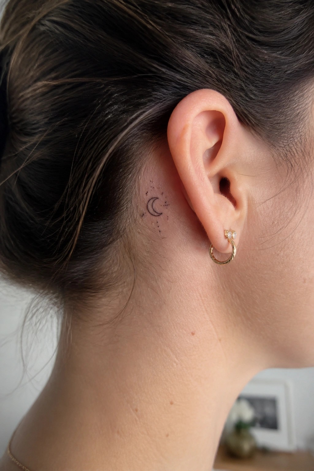Crescent moon and stars tattoo behind ear with gold hoop earring, dark hair pulled back