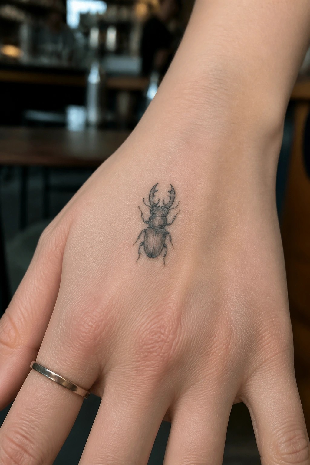 Hand with black stag beetle tattoo and thin metallic ring on finger.