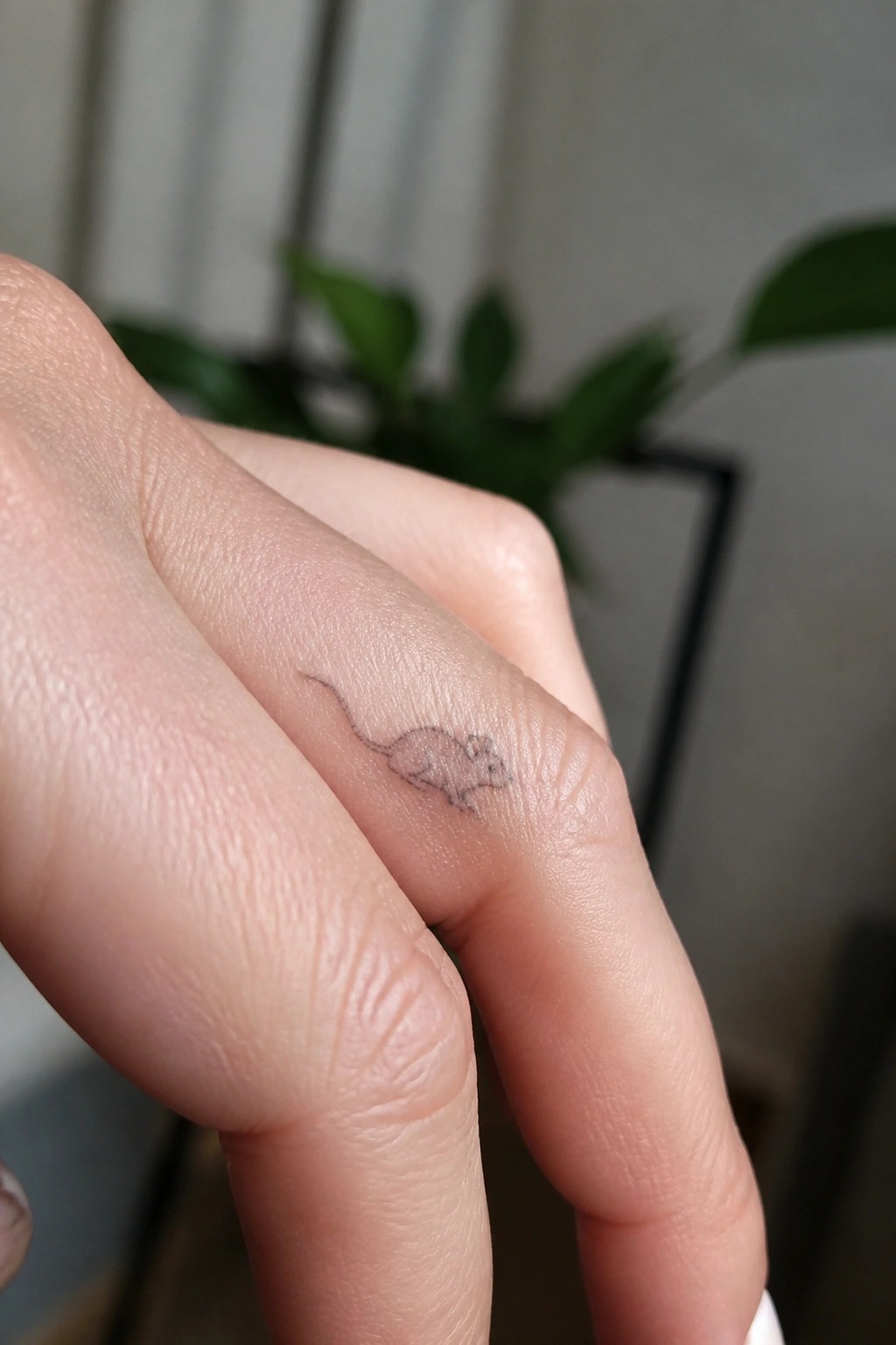 Tiny mouse tattoo on side of finger with blurred green plants behind.