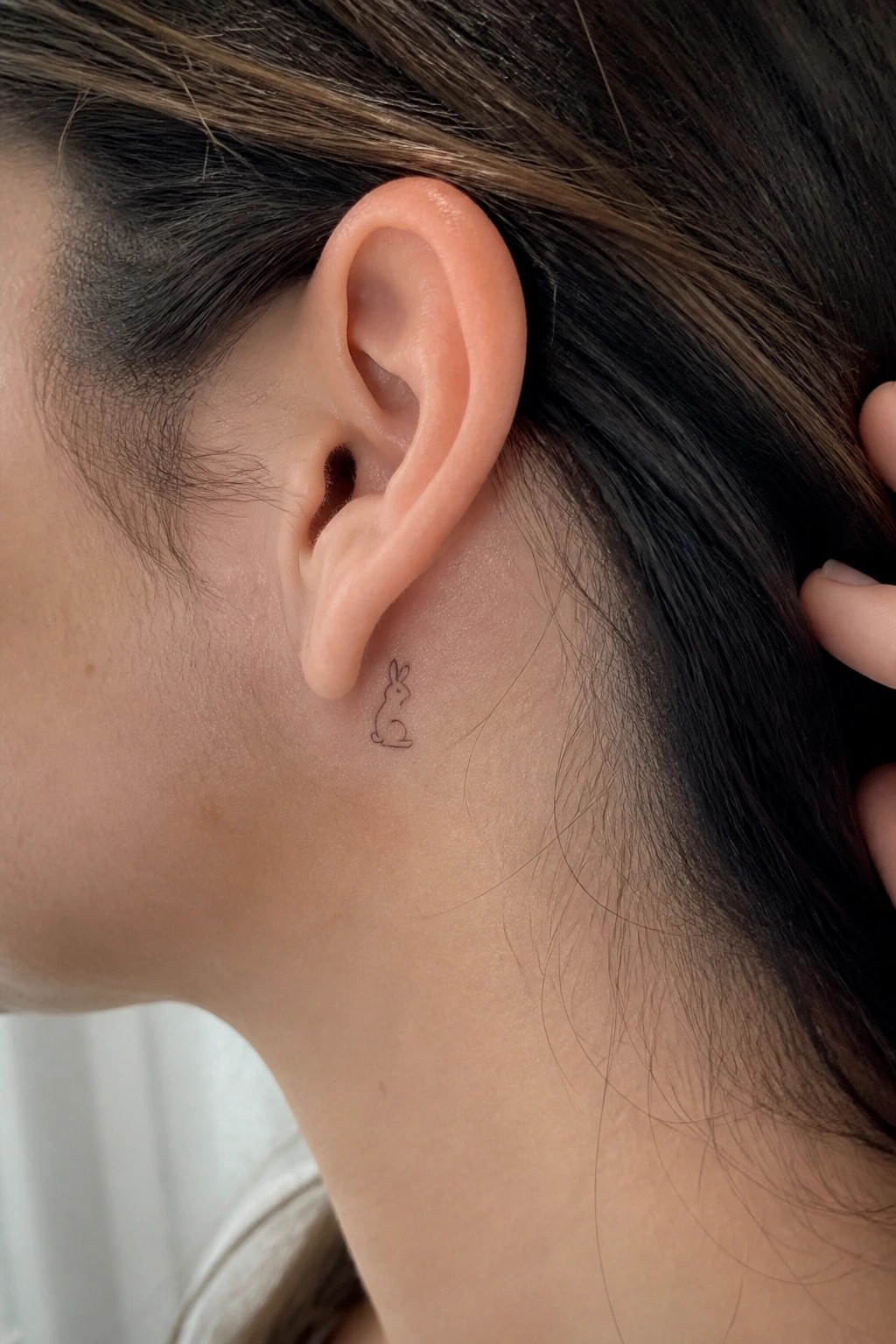 Small rabbit tattoo behind a person's ear on light skin with dark hair.