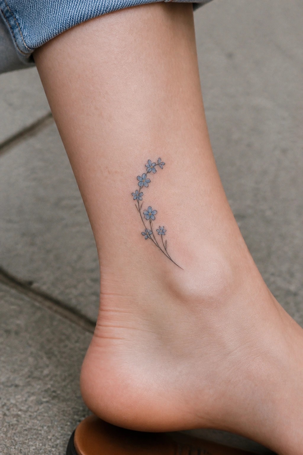 Small blue flowers tattoo on the ankle with a thin curving stem in fine line style