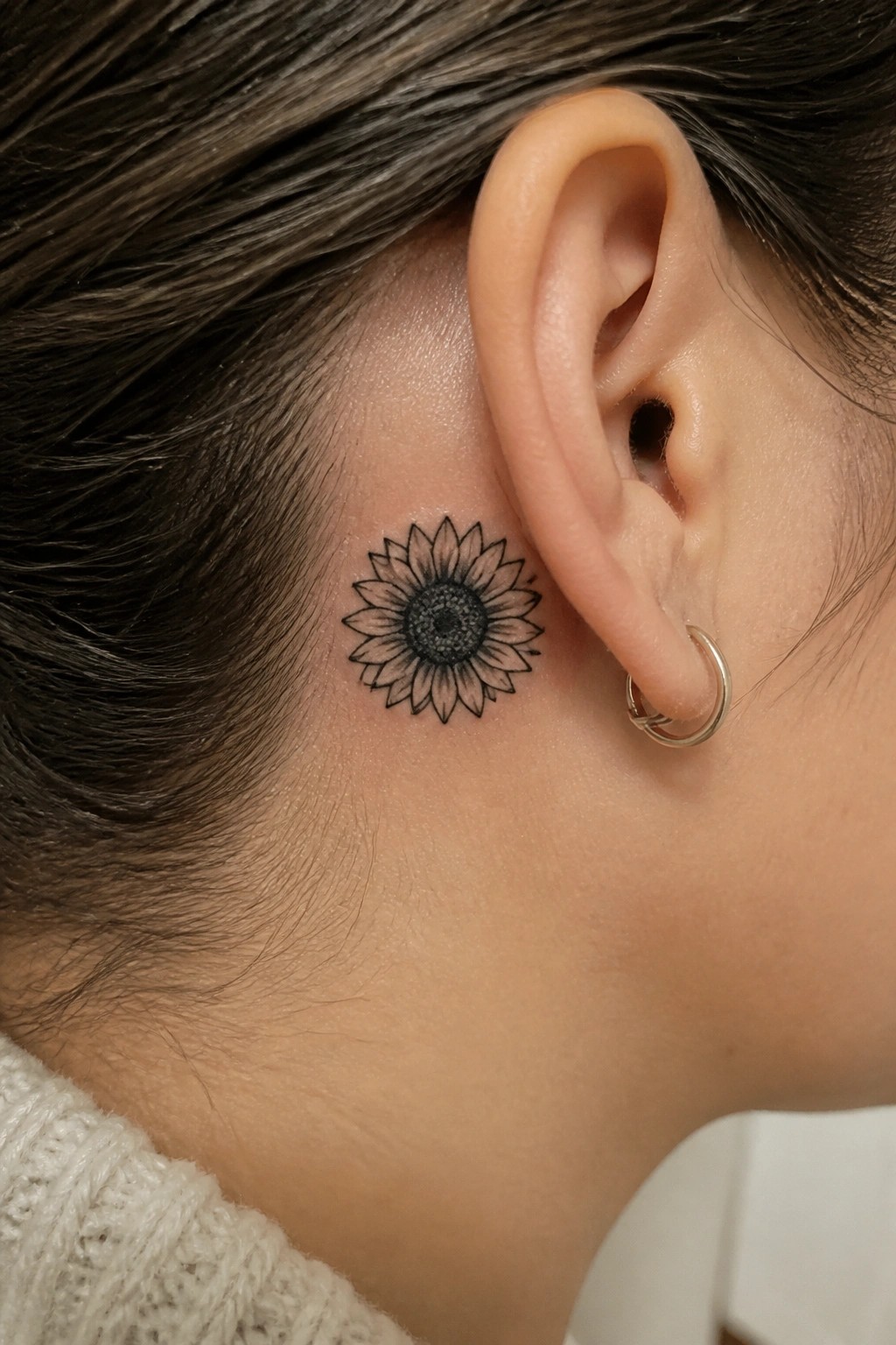 Small black and grey sunflower tattoo placed behind the ear along the hairline on the side of the head.