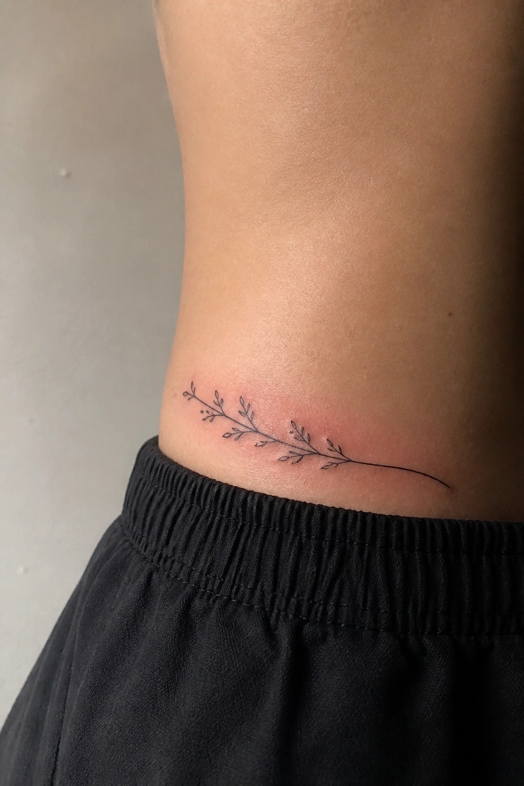 Delicate black leafy branch tattoo on skin above black elastic waistband
