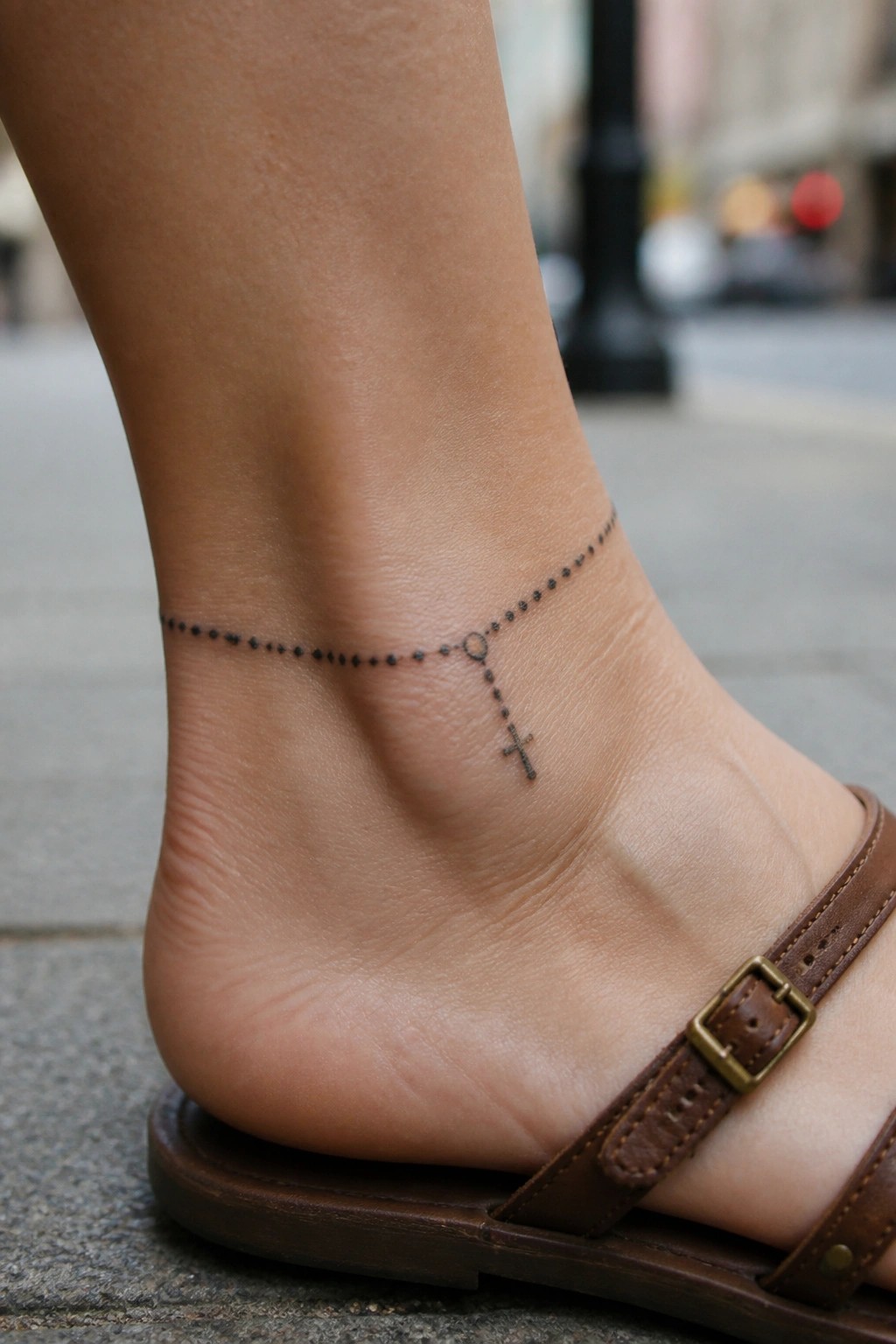Fine dot rosary chain tattoo wrapped around the ankle with a small cross dangling on the side of the foot
