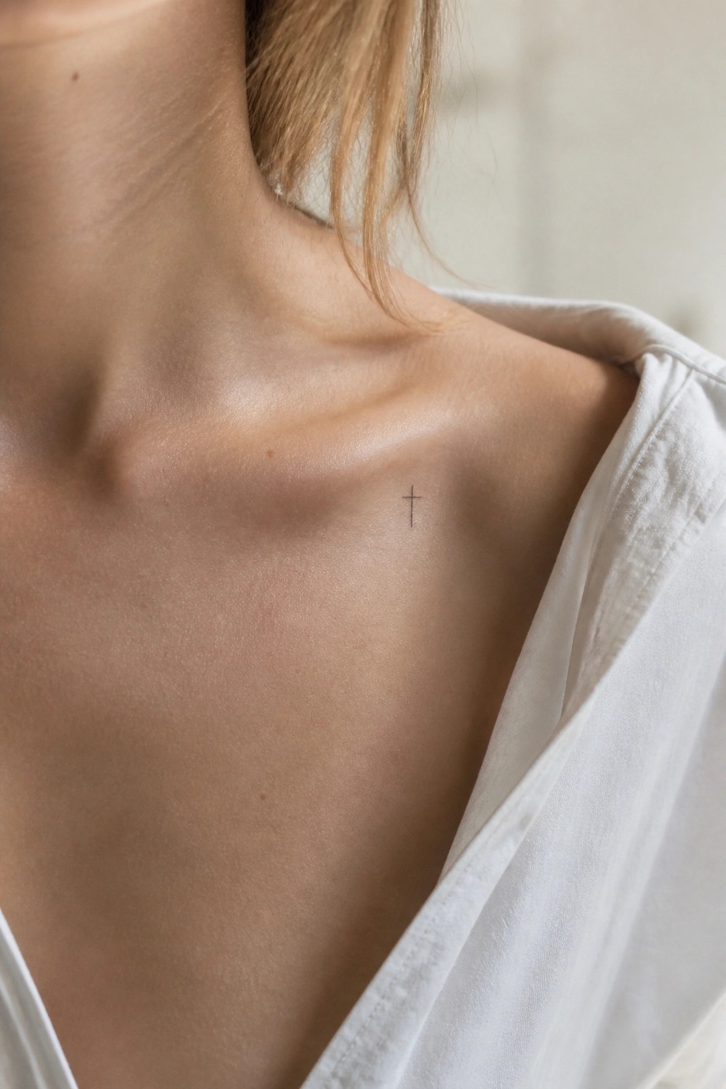 Small cross tattoo on light-skinned shoulder beneath draped white shirt fabric.