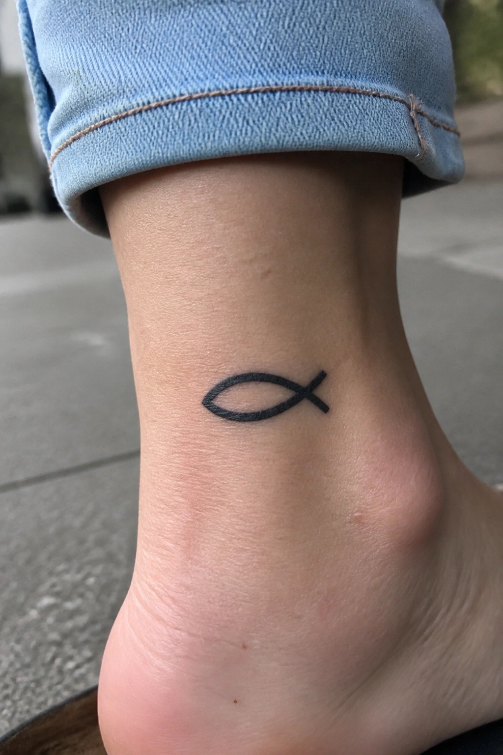 Black fish tattoo on ankle below rolled cuff of blue jeans