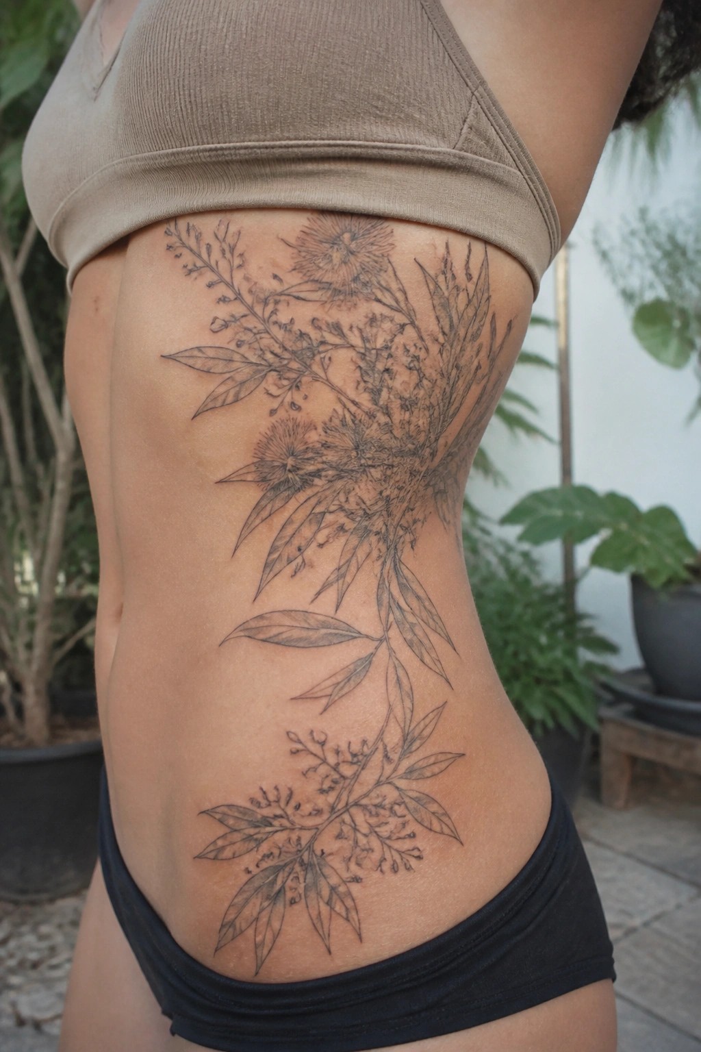 Fine line floral tattoo on side torso under beige crop top and black bottoms