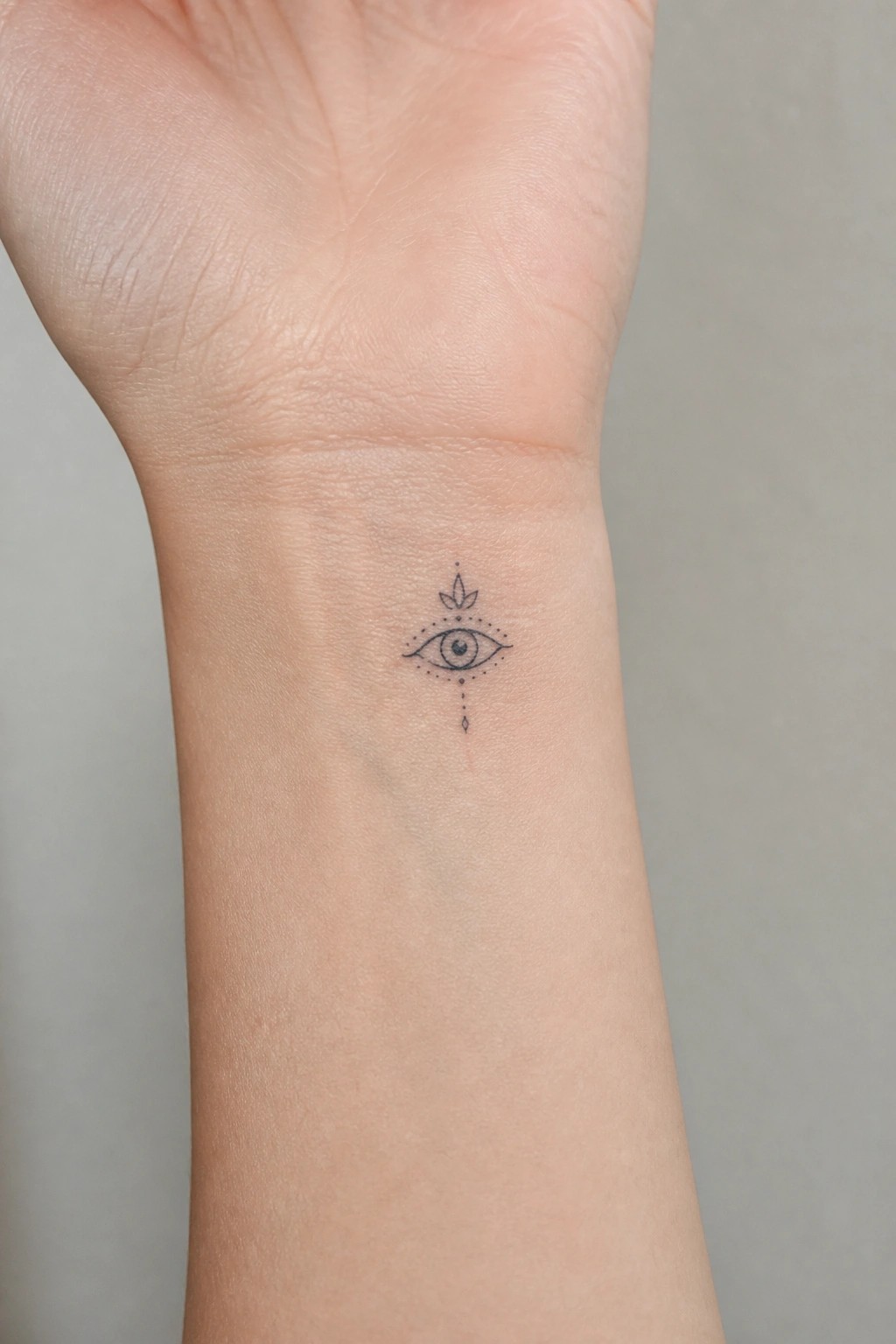 Small fine line eye tattoo centered on the inner wrist with dotted accents and a minimal leaf detail above.
