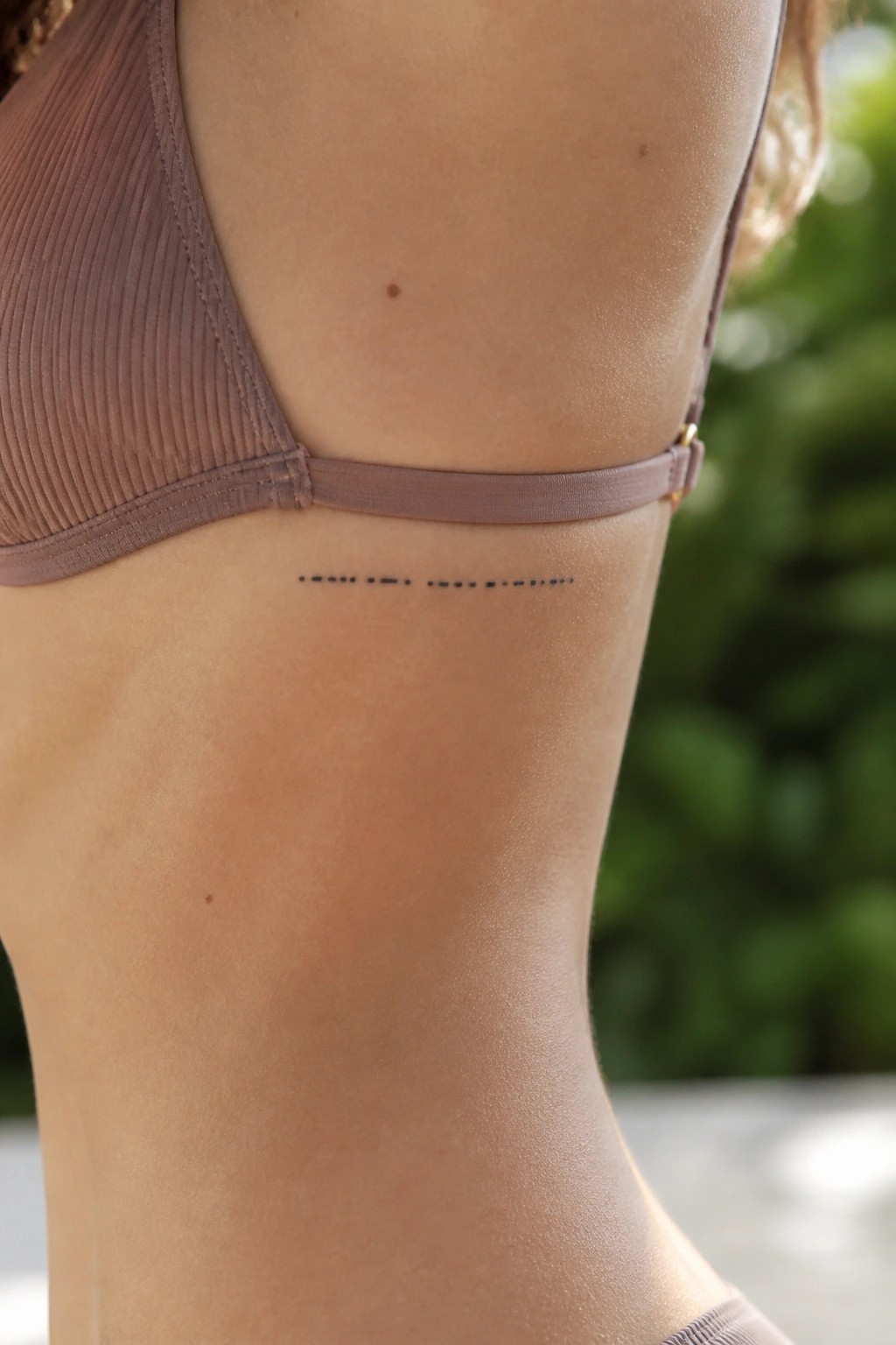 Woman's side in ribbed mauve bikini top with small dashed-line tattoo on ribs