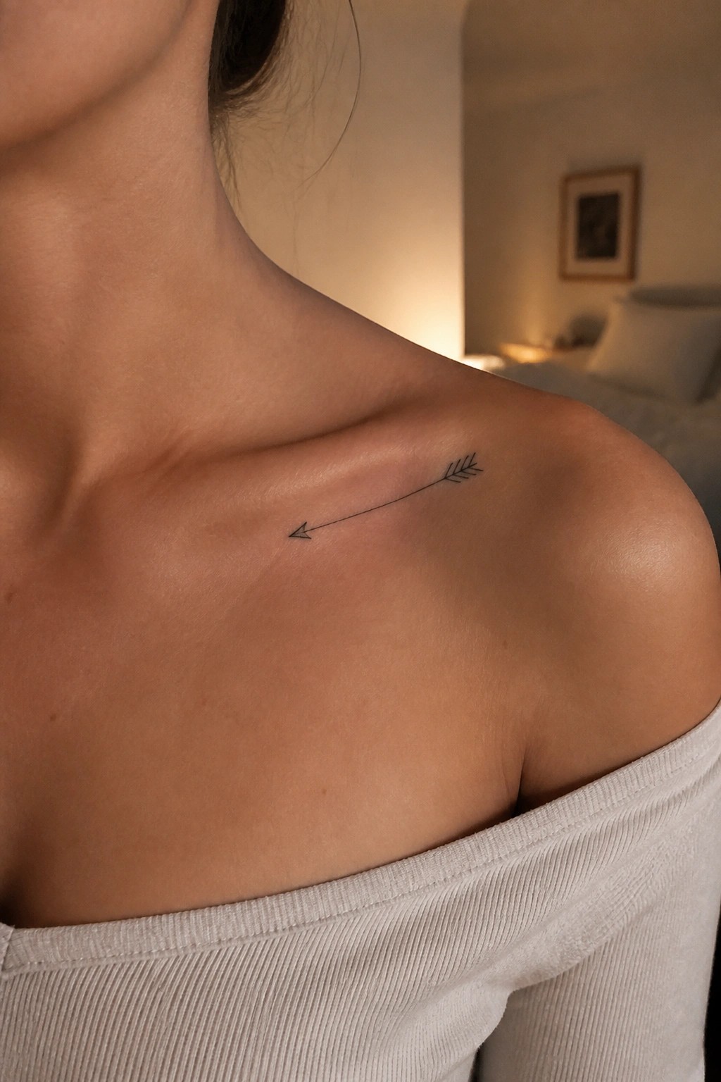 Woman's shoulder with black arrow tattoo above off-shoulder ribbed beige top in bedroom