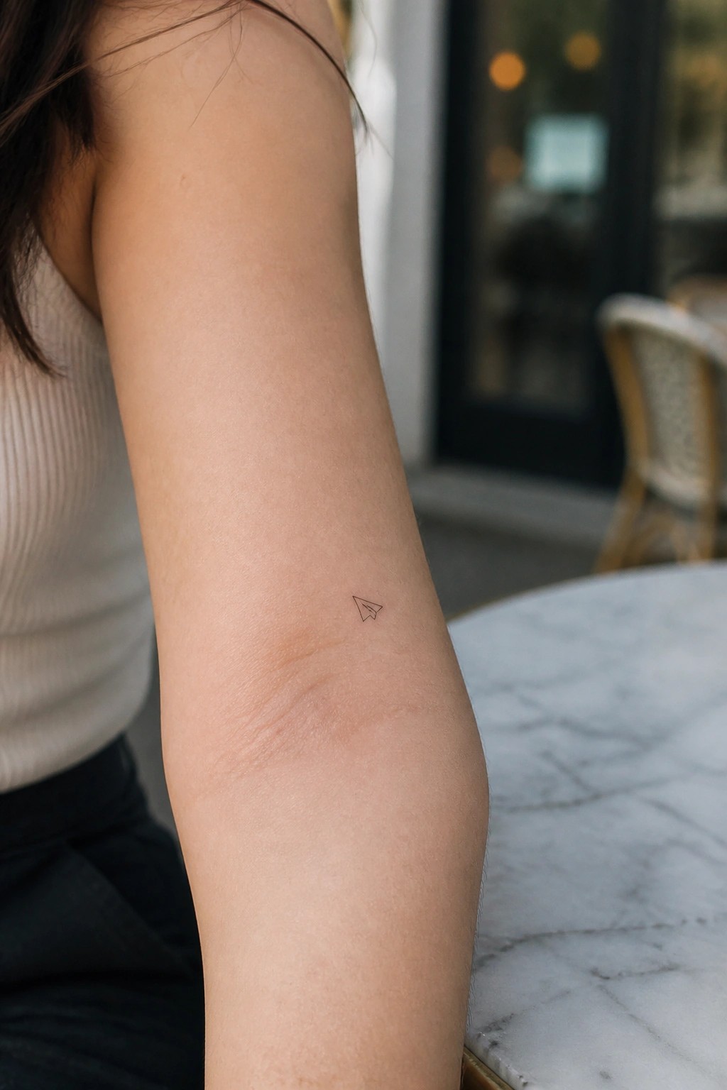 Small paper airplane tattoo on upper arm in white ribbed tank top on marble table