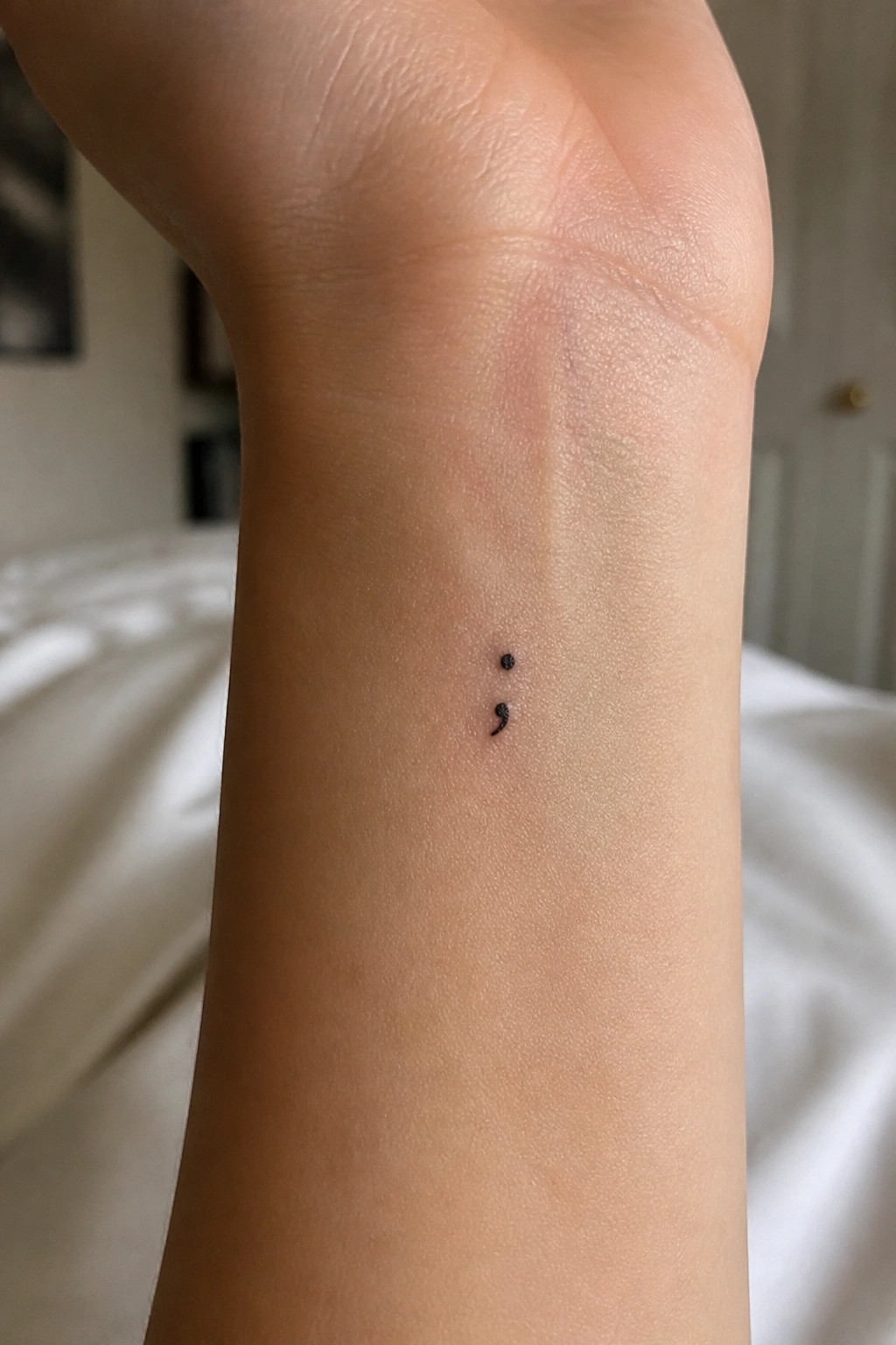 Small black semicolon tattoo on inner wrist of light-skinned arm.