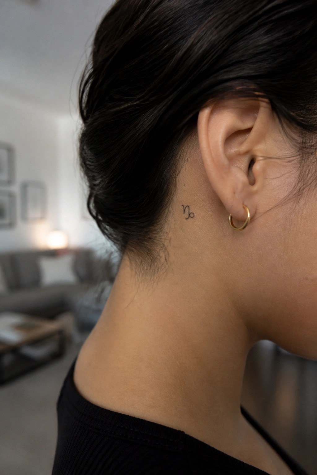 Woman's neck with Capricorn tattoo, gold hoop earring, dark bun, black top