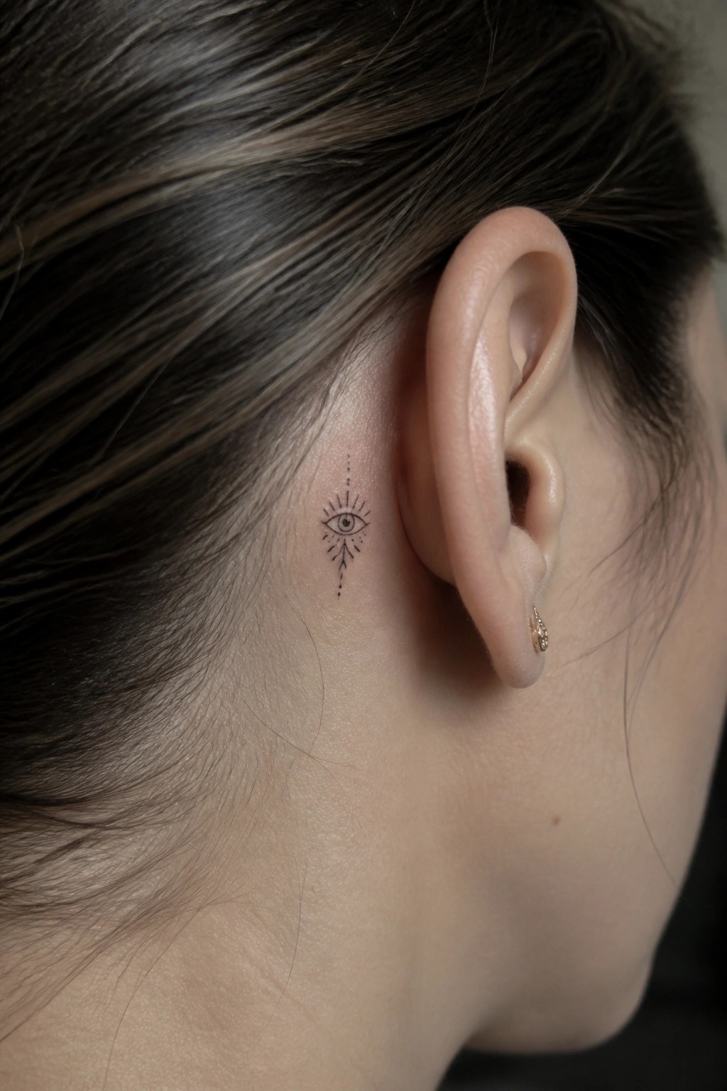 Small eye tattoo behind ear with tiny gold earring in lobe