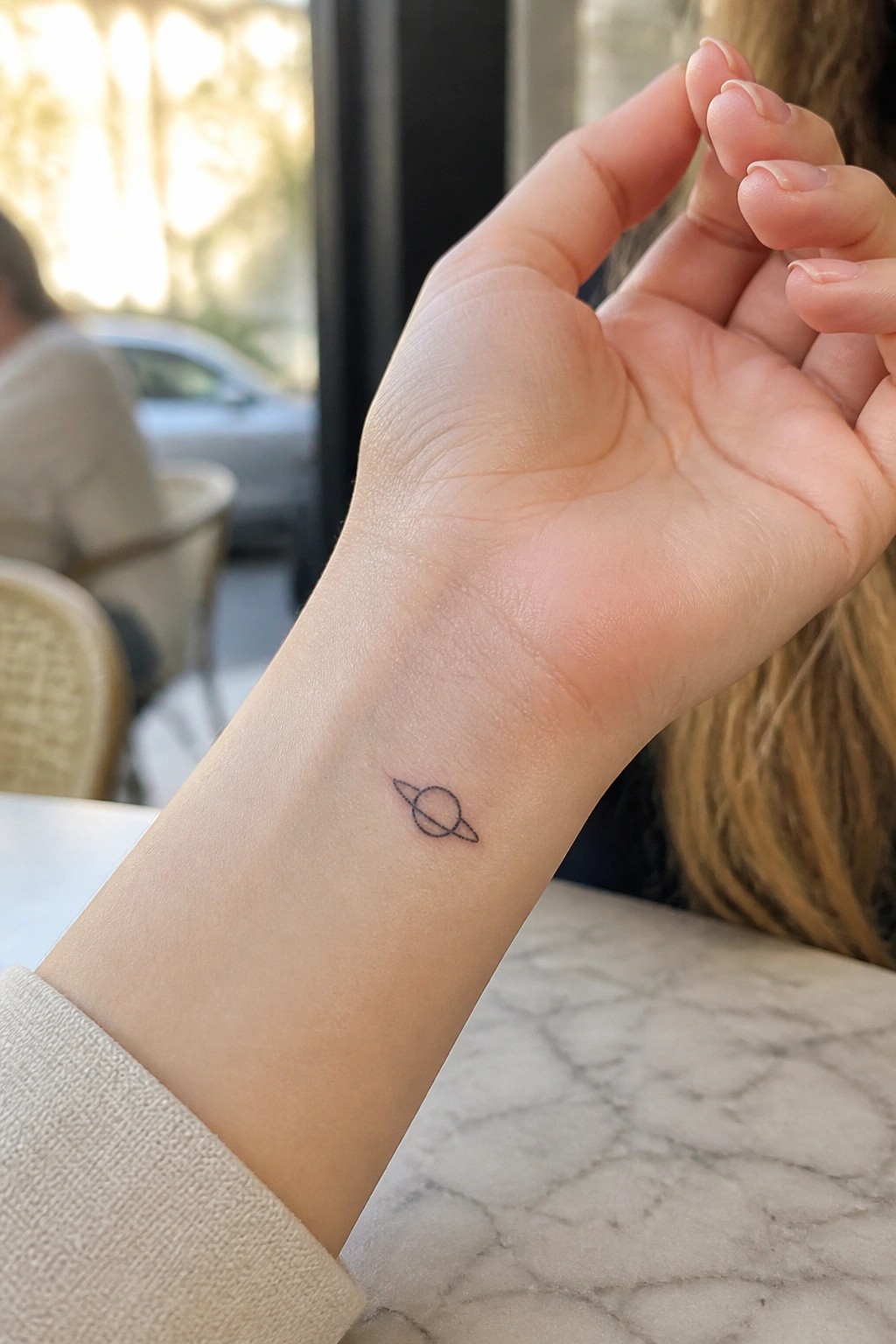 Small Saturn tattoo on wrist above marble table in bright cafe setting.