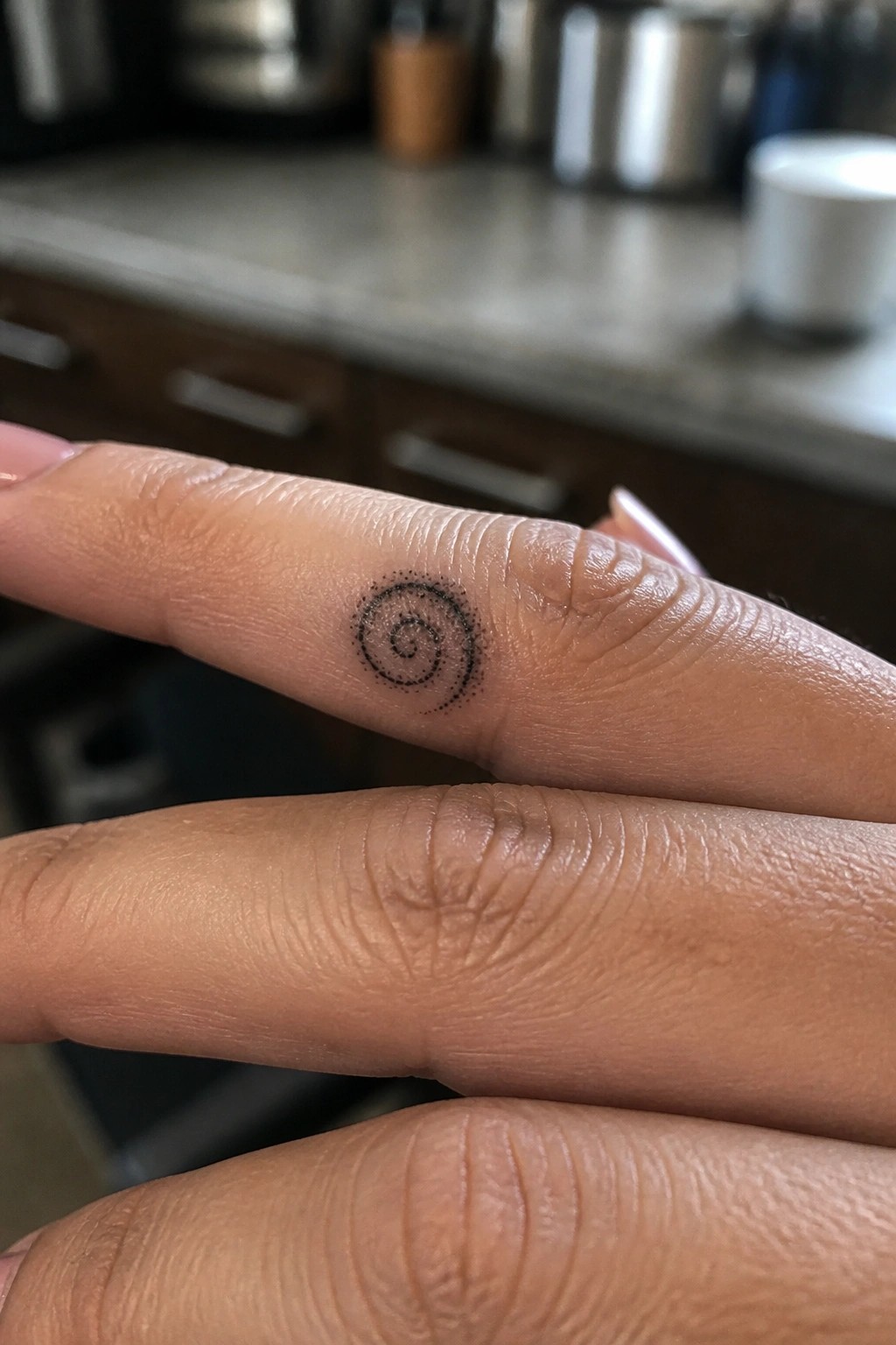 Finger with black dotted spiral tattoo and manicured nails, blurred kitchen background