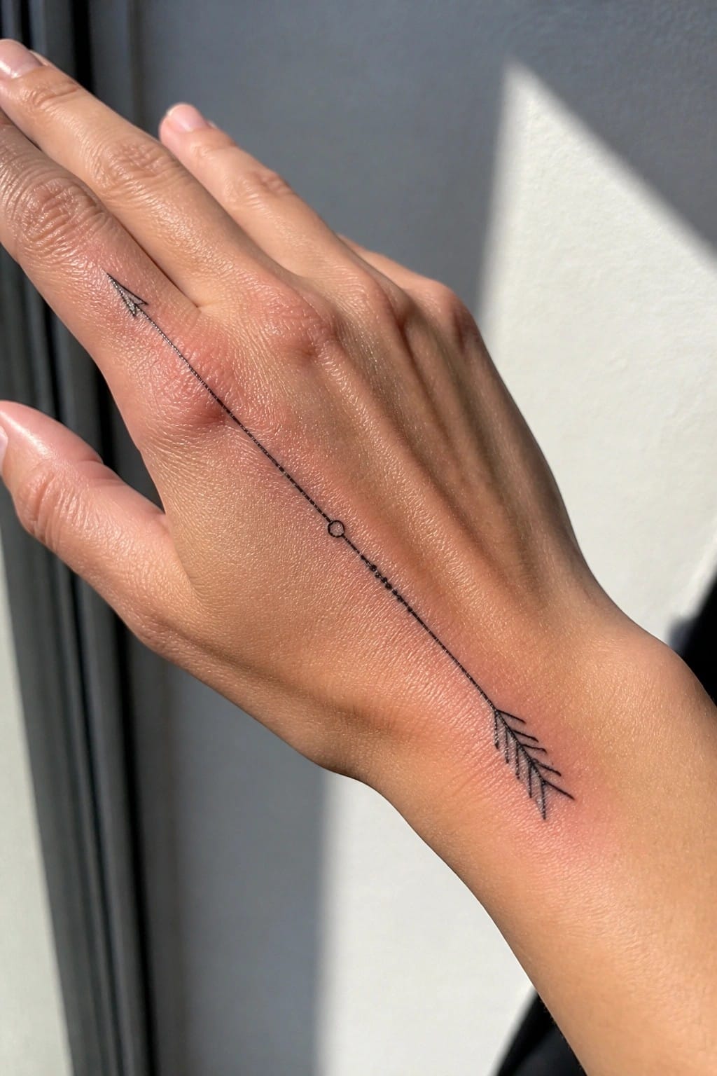 Fine line arrow tattoo spanning the back of a hand from near the index finger to the wrist area with a pointed tip and feathered end.