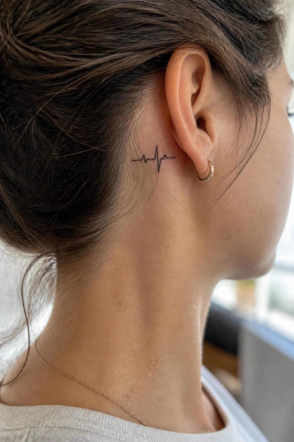 Woman's neck with heartbeat tattoo behind ear, gold hoop earring, and thin necklace