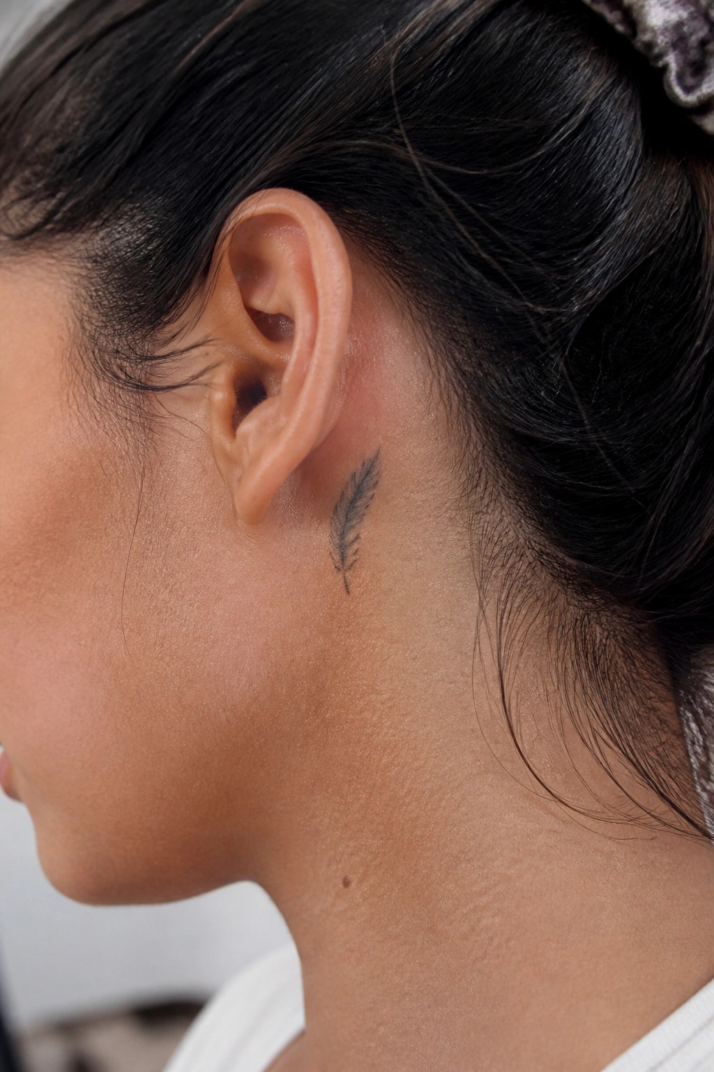 Small black feather tattoo placed behind the ear on the side of the neck