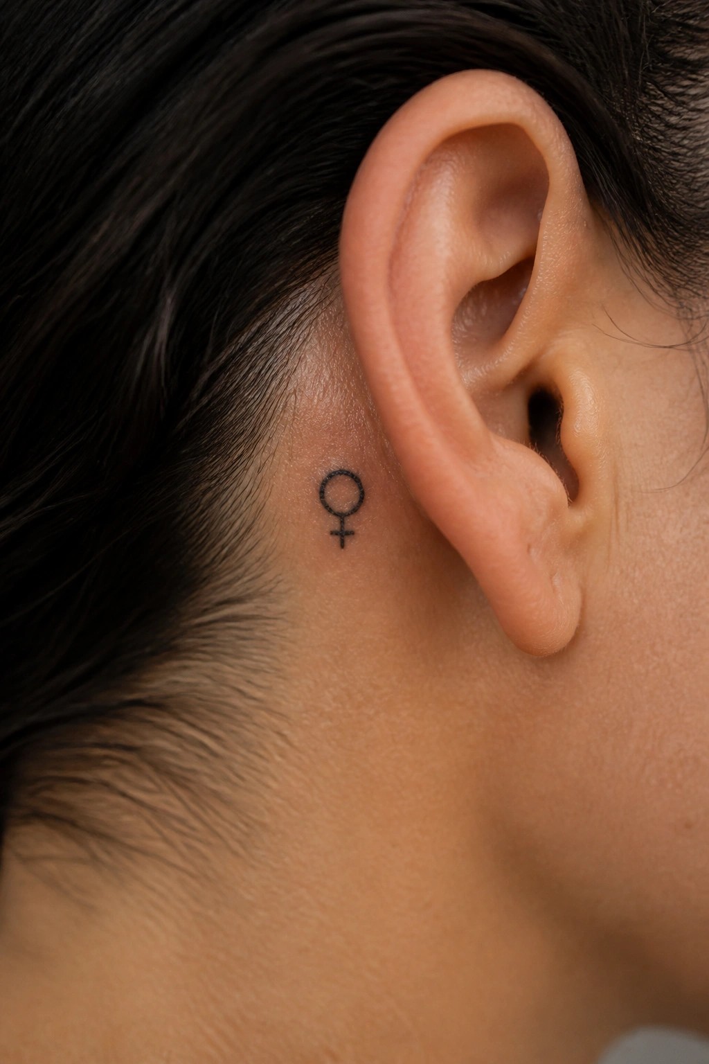 Small black female gender symbol tattoo placed just behind the ear on bare skin near the hairline