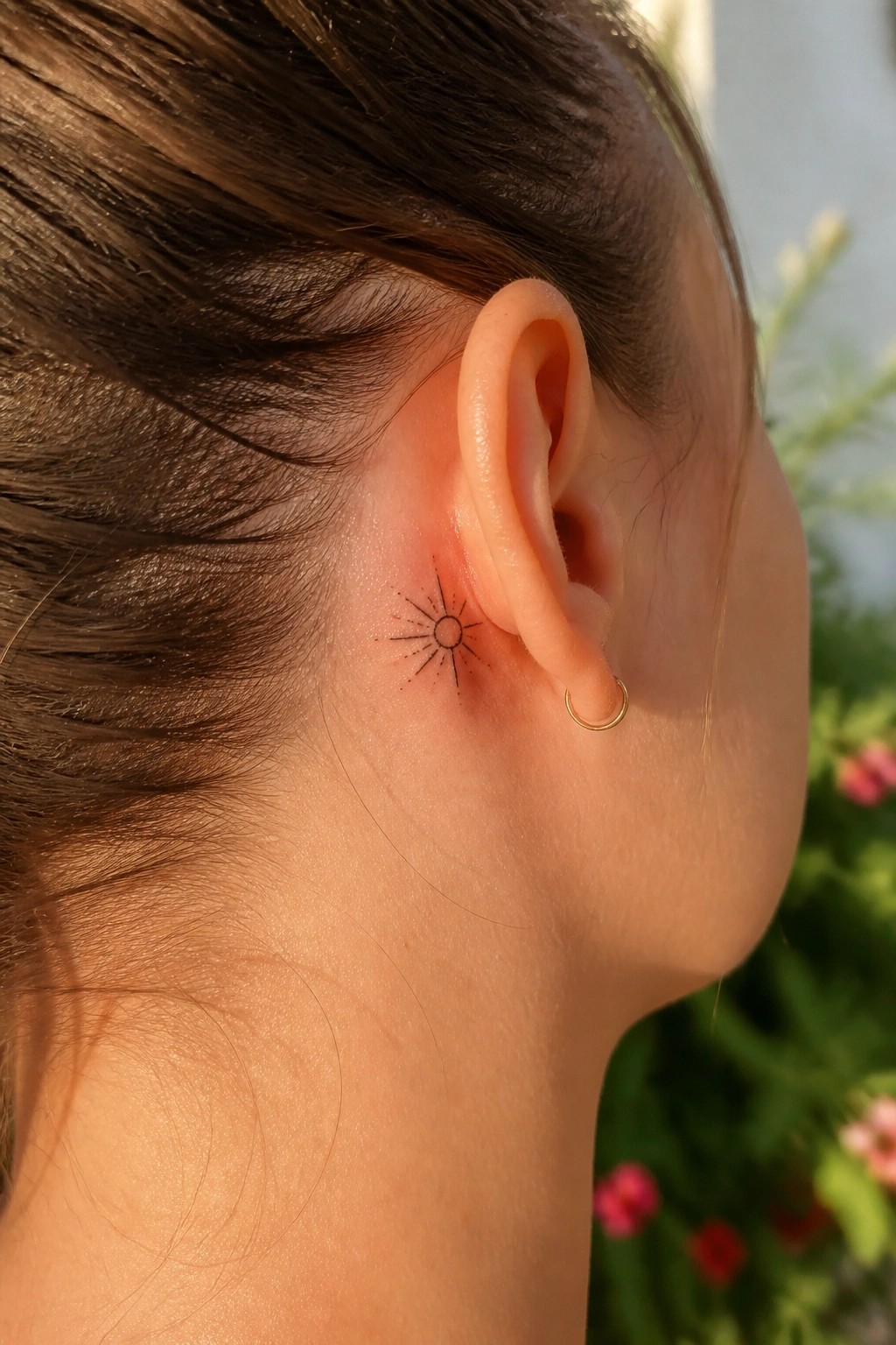 Sun tattoo behind ear with small gold hoop earring on tanned skin