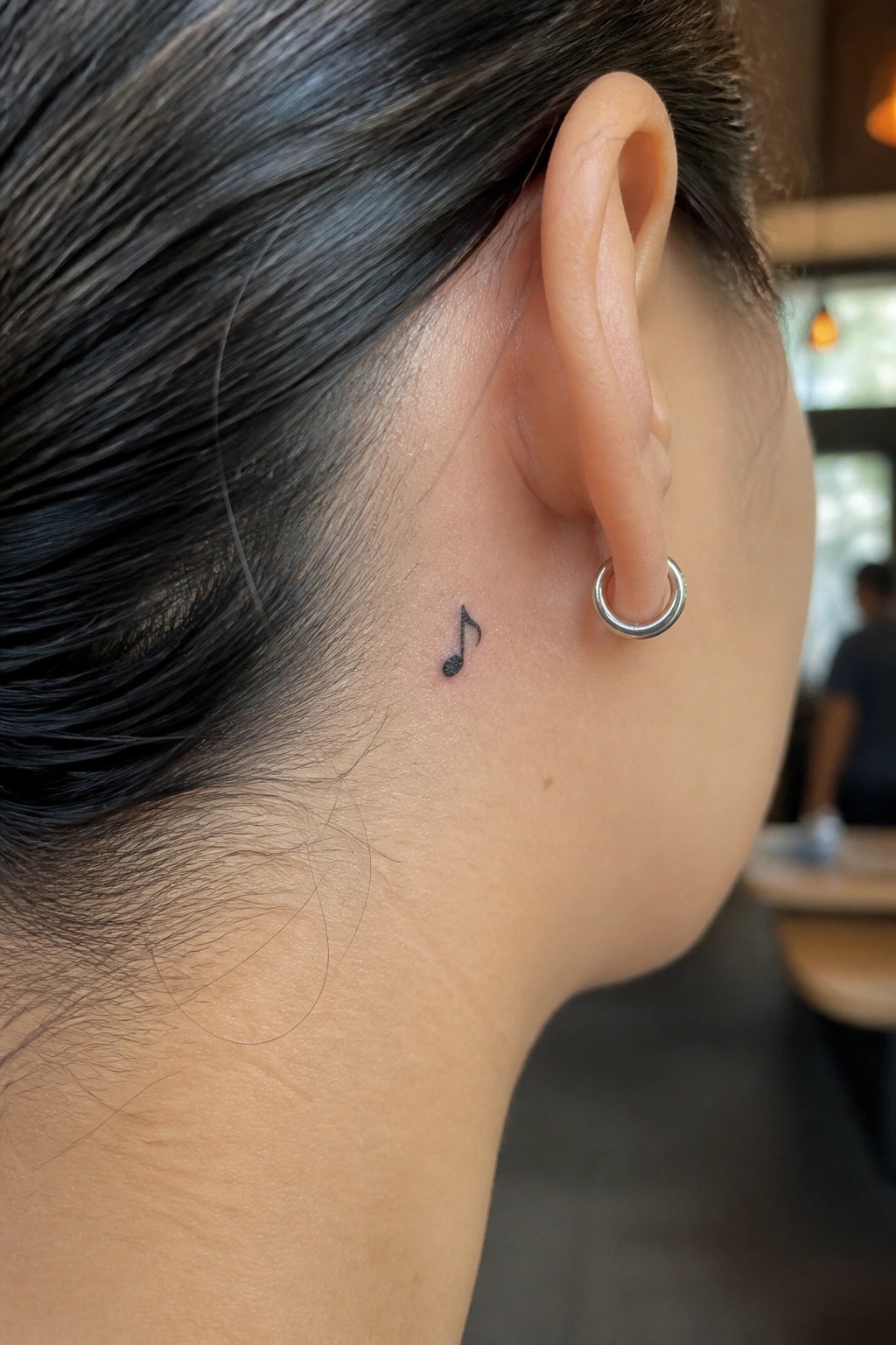 Small black music note tattoo behind the ear next to a silver hoop earring