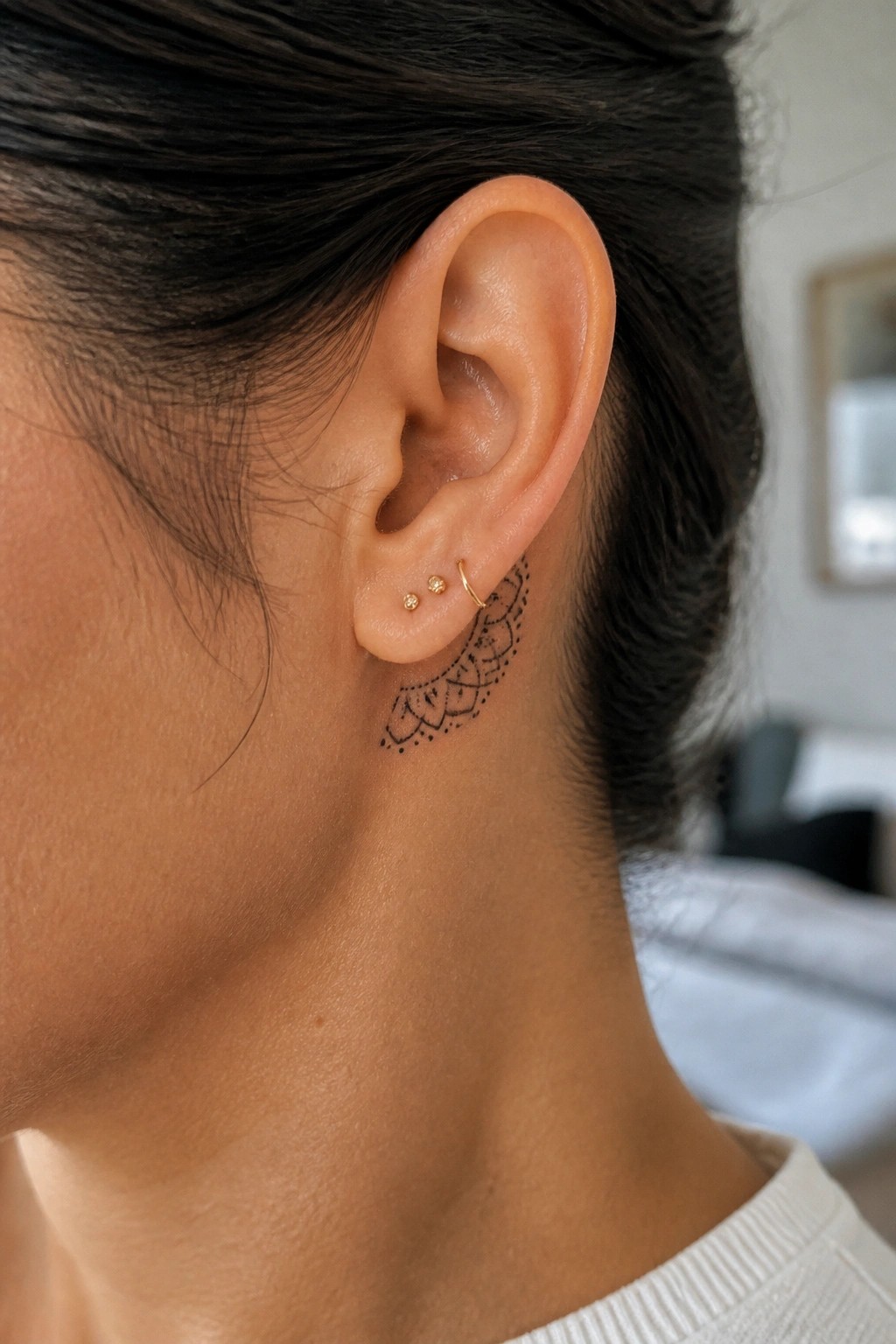 Close up of a small black mandala tattoo placed behind the ear on the neck with fine line details and three small gold earrings nearby.