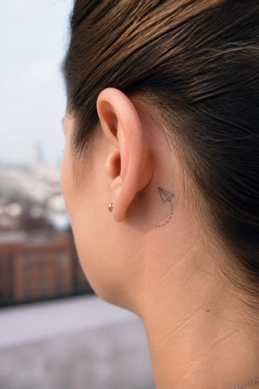 Close-up of ear with paper airplane tattoo, dotted line, and gold stud earring.
