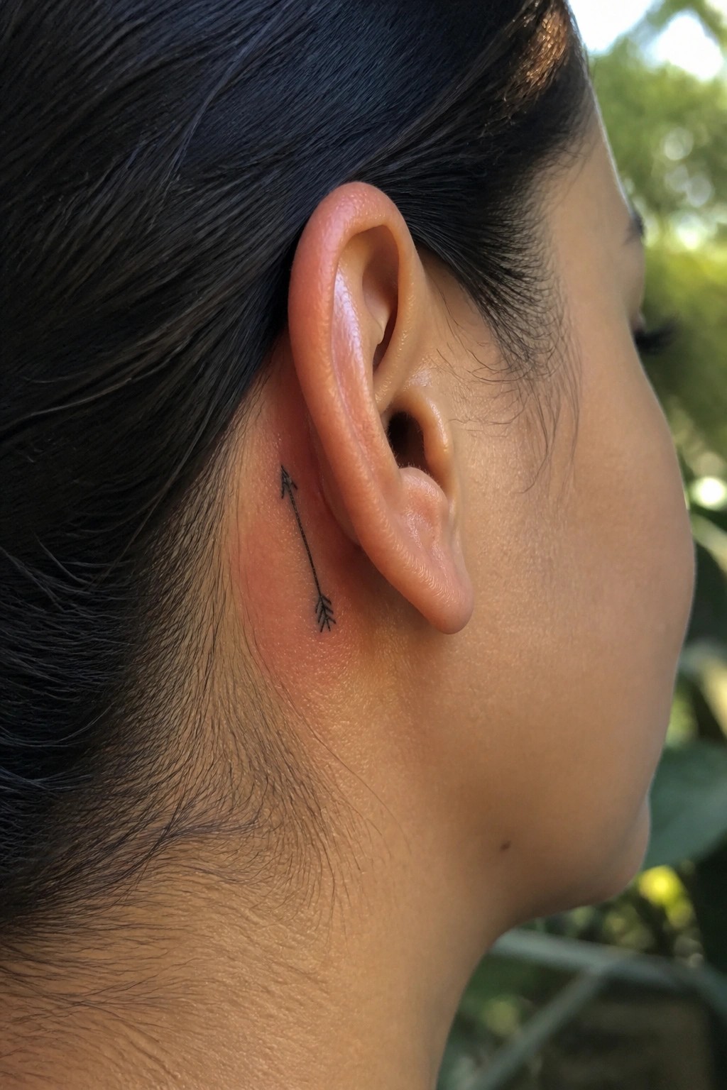 Minimalist black arrow tattoo placed behind a woman's ear, pointing upward with a feathered base.