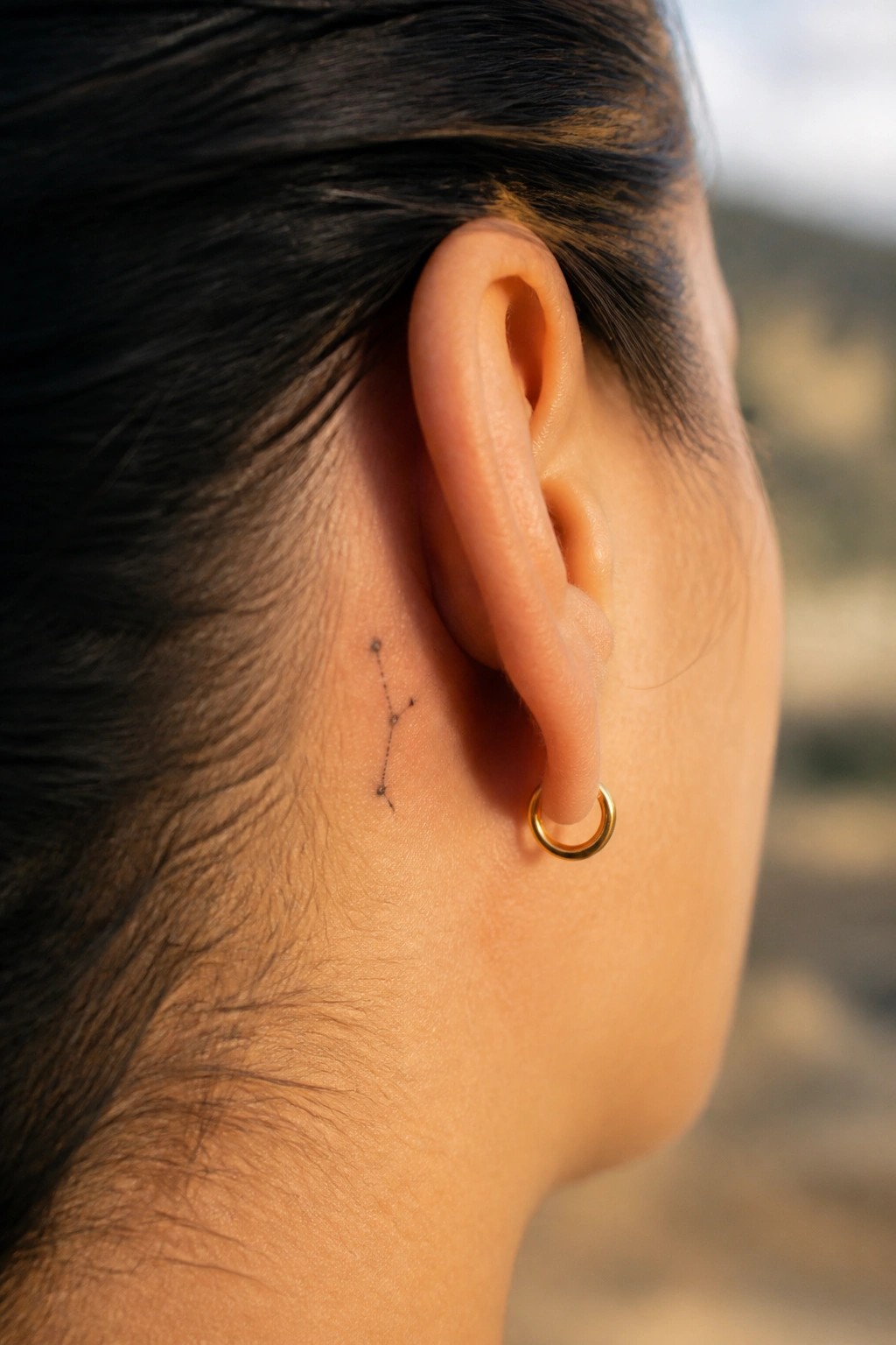 Small constellation tattoo made of dots and lines placed behind the ear next to a gold hoop earring.