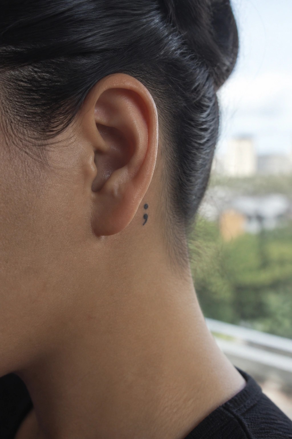 Close-up of ear with semicolon tattoo behind it, dark hair in bun, black shirt