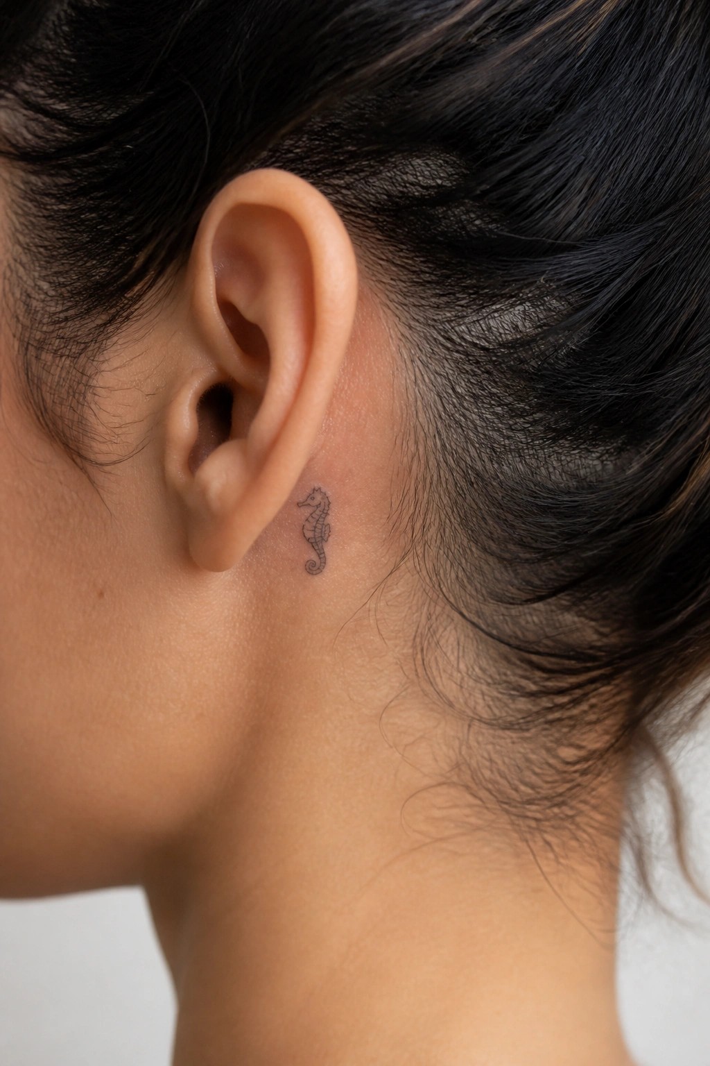 Small black seahorse tattoo behind a person's ear with dark hair.