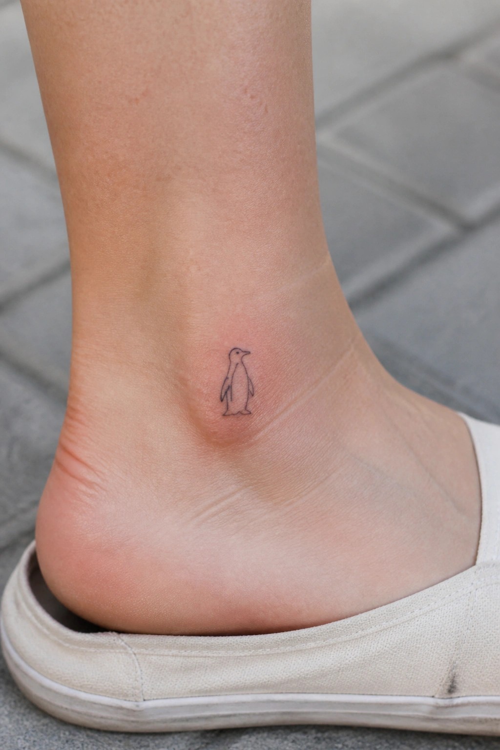 Minimalist black line drawing of a small penguin tattoo on the ankle above a white shoe