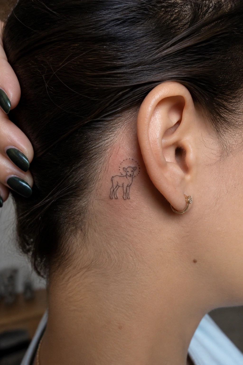 Lamb tattoo with halo behind ear beside gold hoop earring, green nails visible